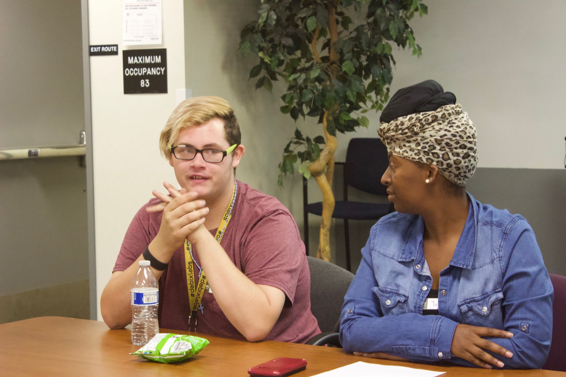 Two people seated at a table, one speaking while the other listens in a meeting room.
