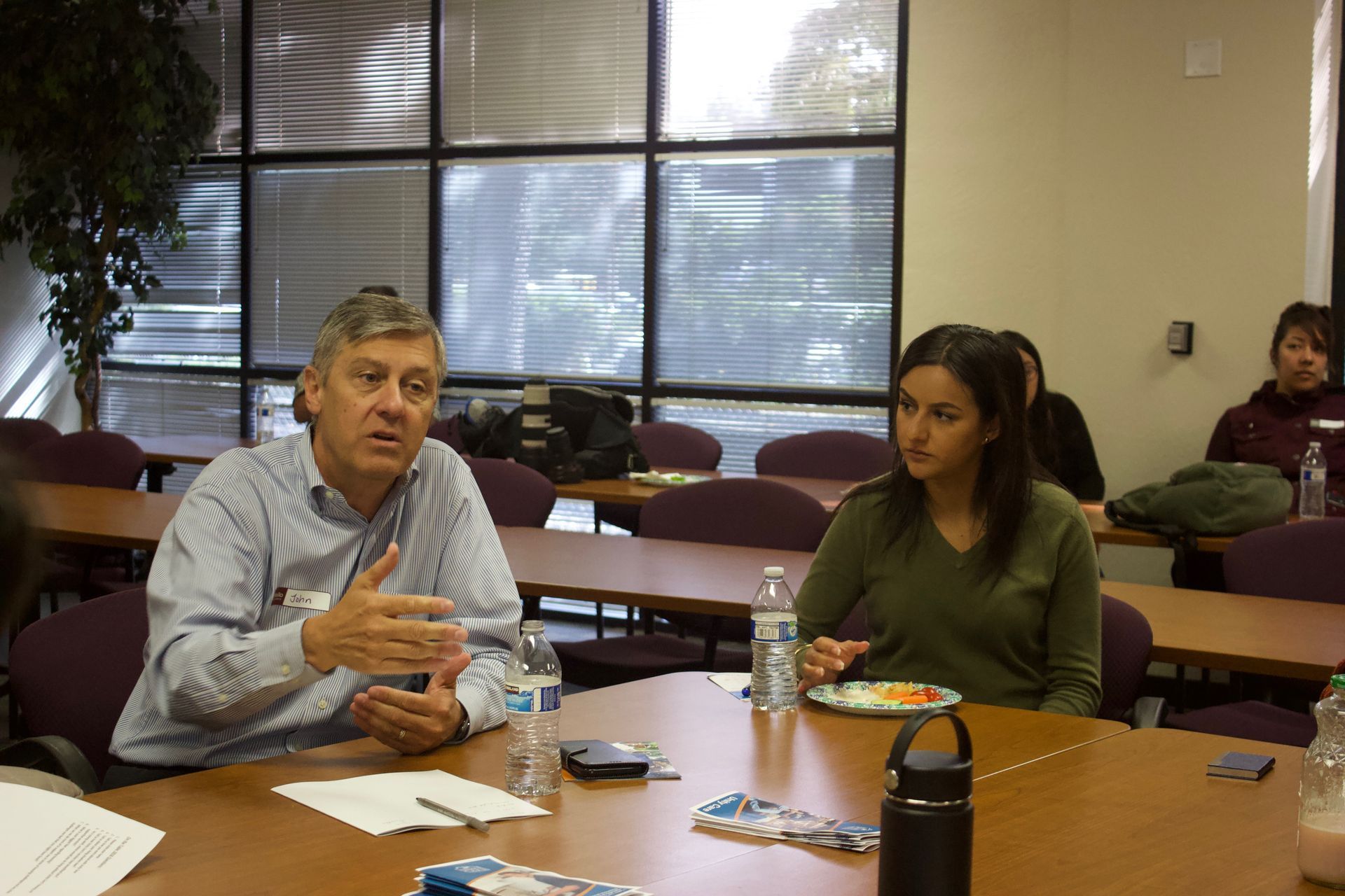 Two people seated at a conference table in a meeting room, with papers and drinks in front of them.