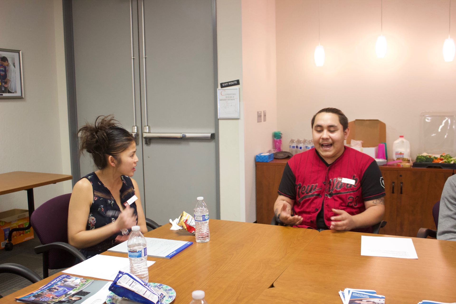 Two people talking at a conference table in a meeting room, with papers and water bottles.