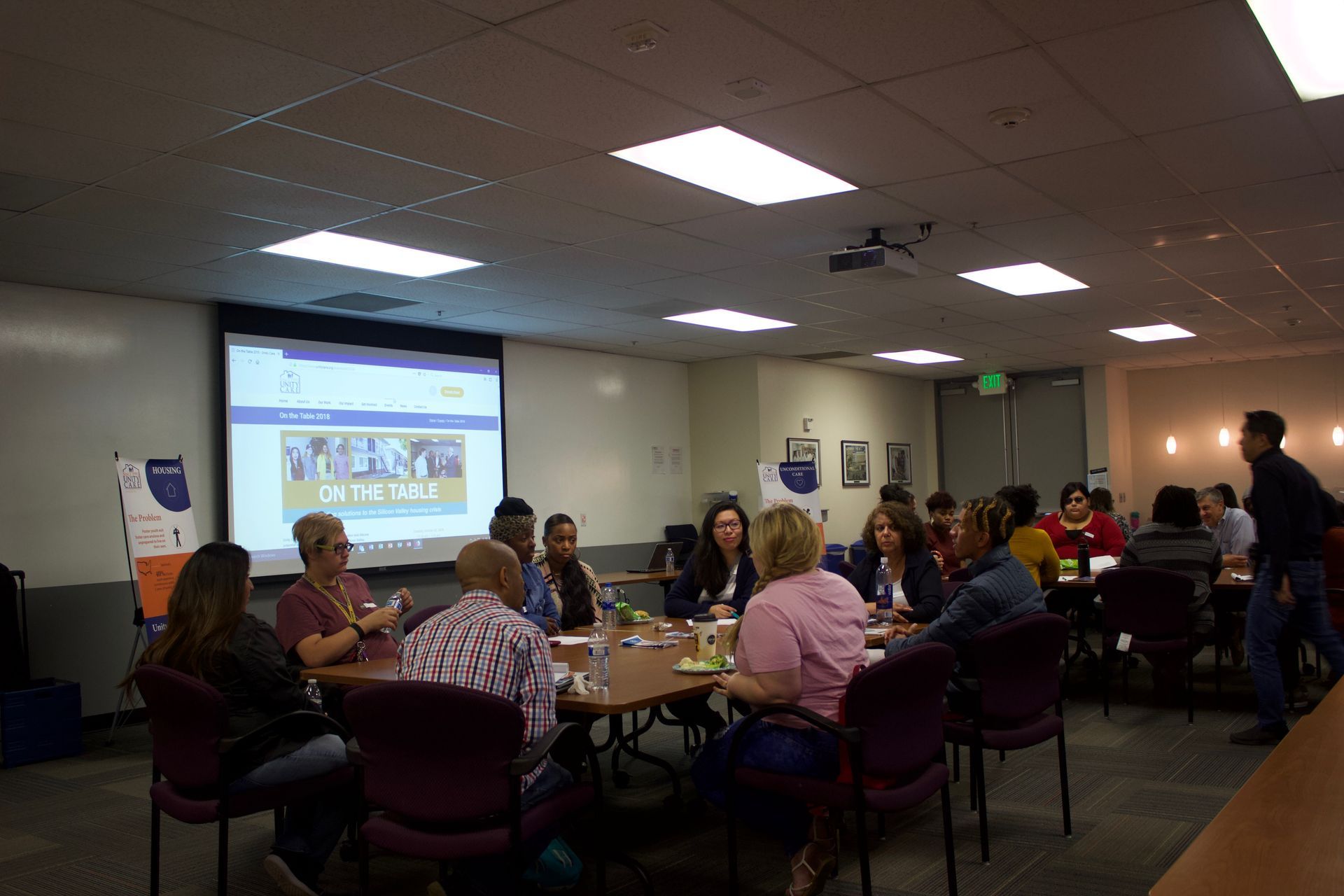 People seated around tables in a meeting room watching a presentation on a projected screen