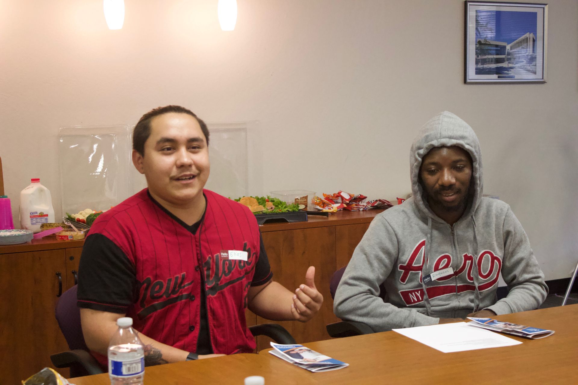 Two people sit at a table in a room, with papers and a water bottle in front of them.