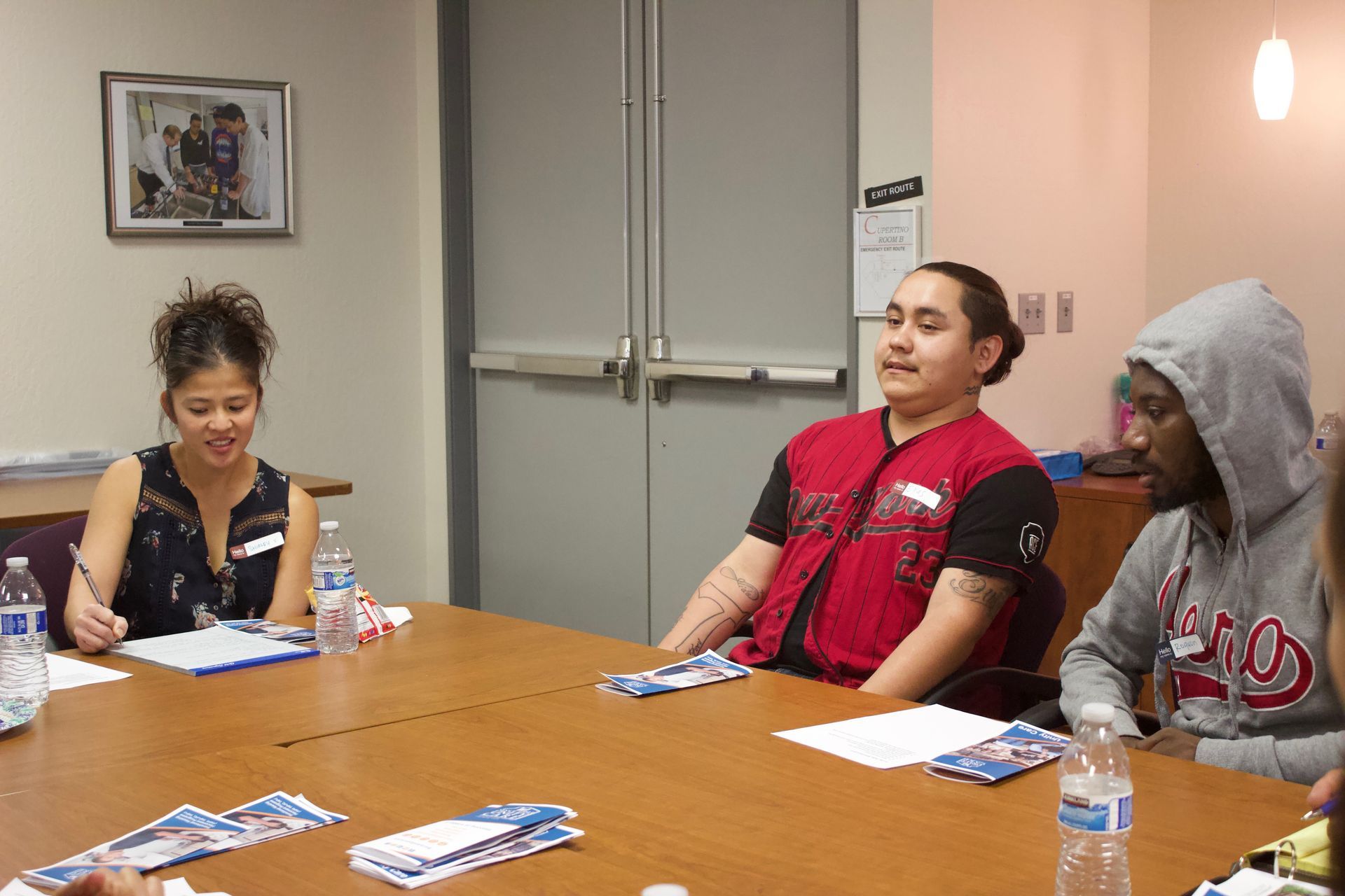 Three people sit around a table in a meeting room with papers, water bottles, and snacks.