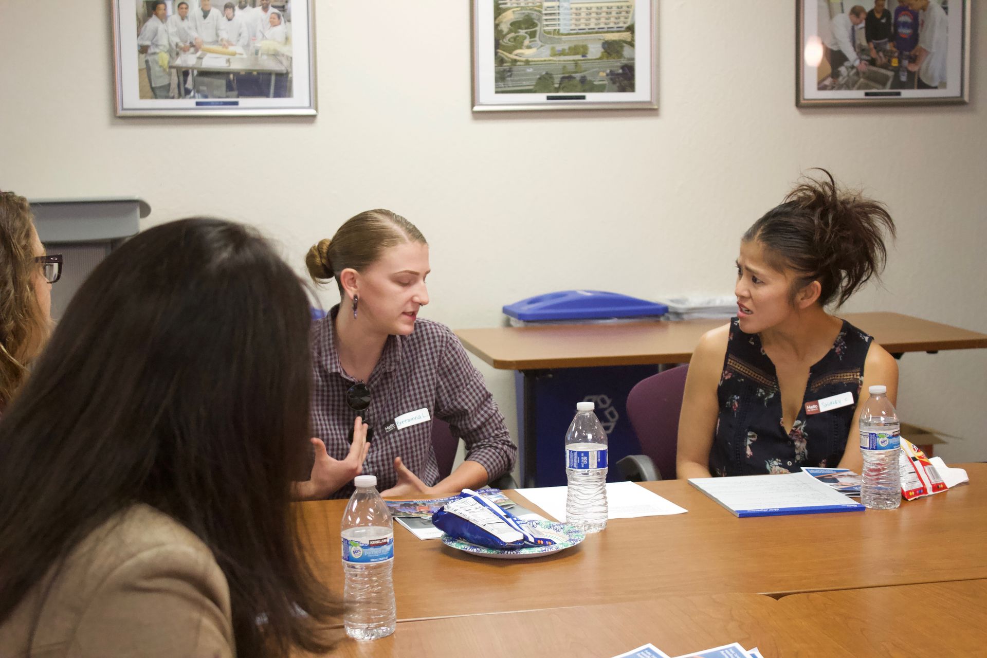 Three women sit at a table in a meeting room, talking with water bottles and papers in front of them.