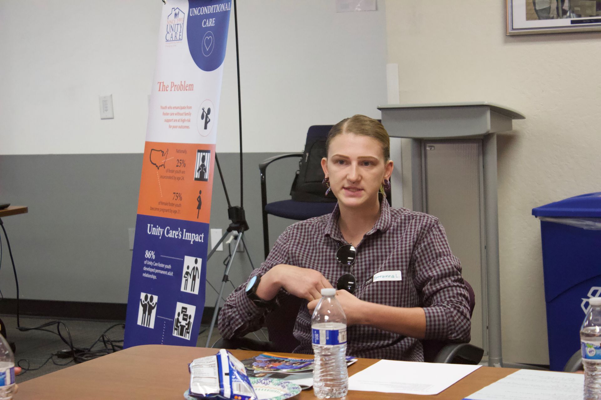 Person seated at a table speaking in a classroom or meeting room, with a banner and water bottle nearby