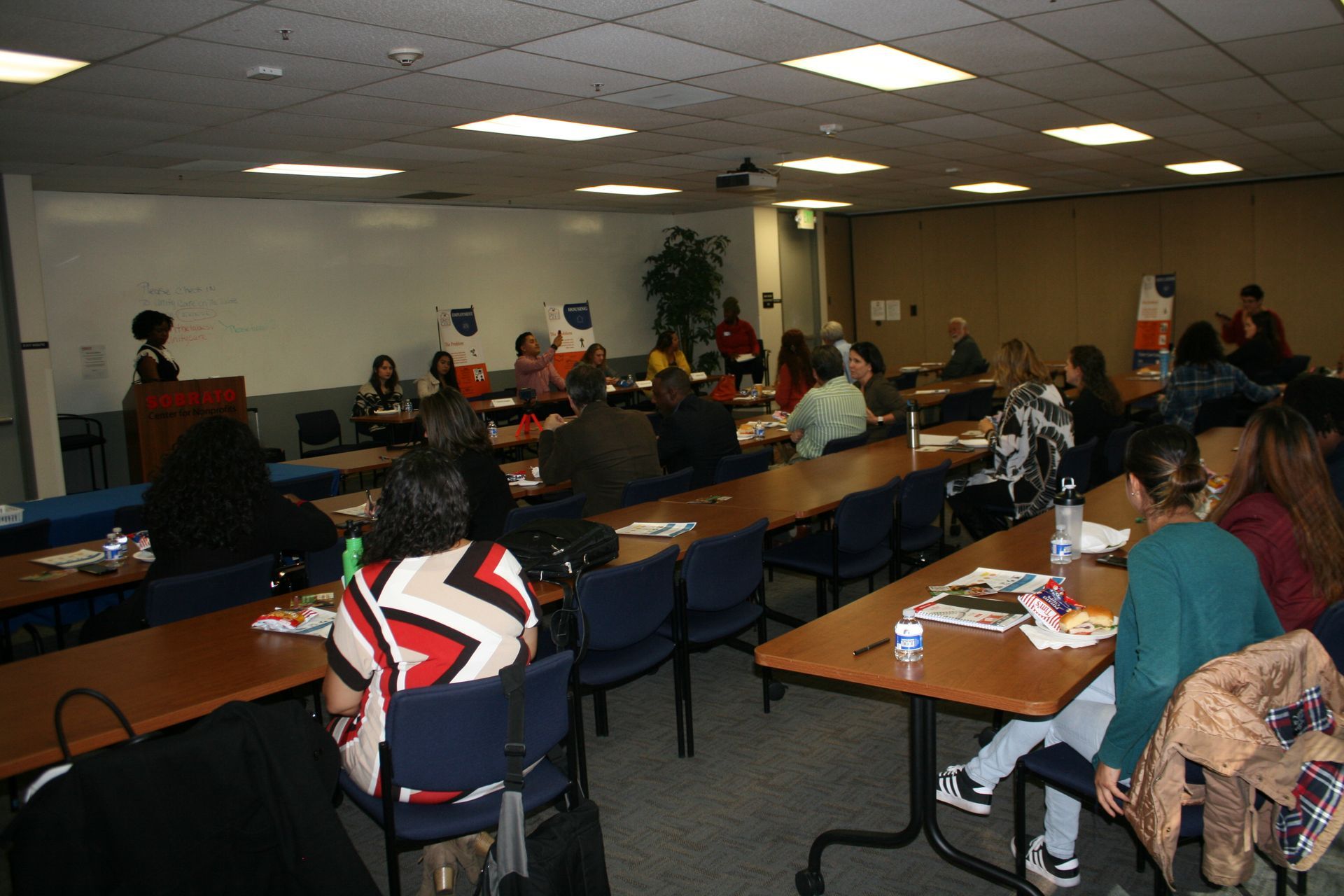 Conference room meeting with people seated at tables, facing a presentation at the front.