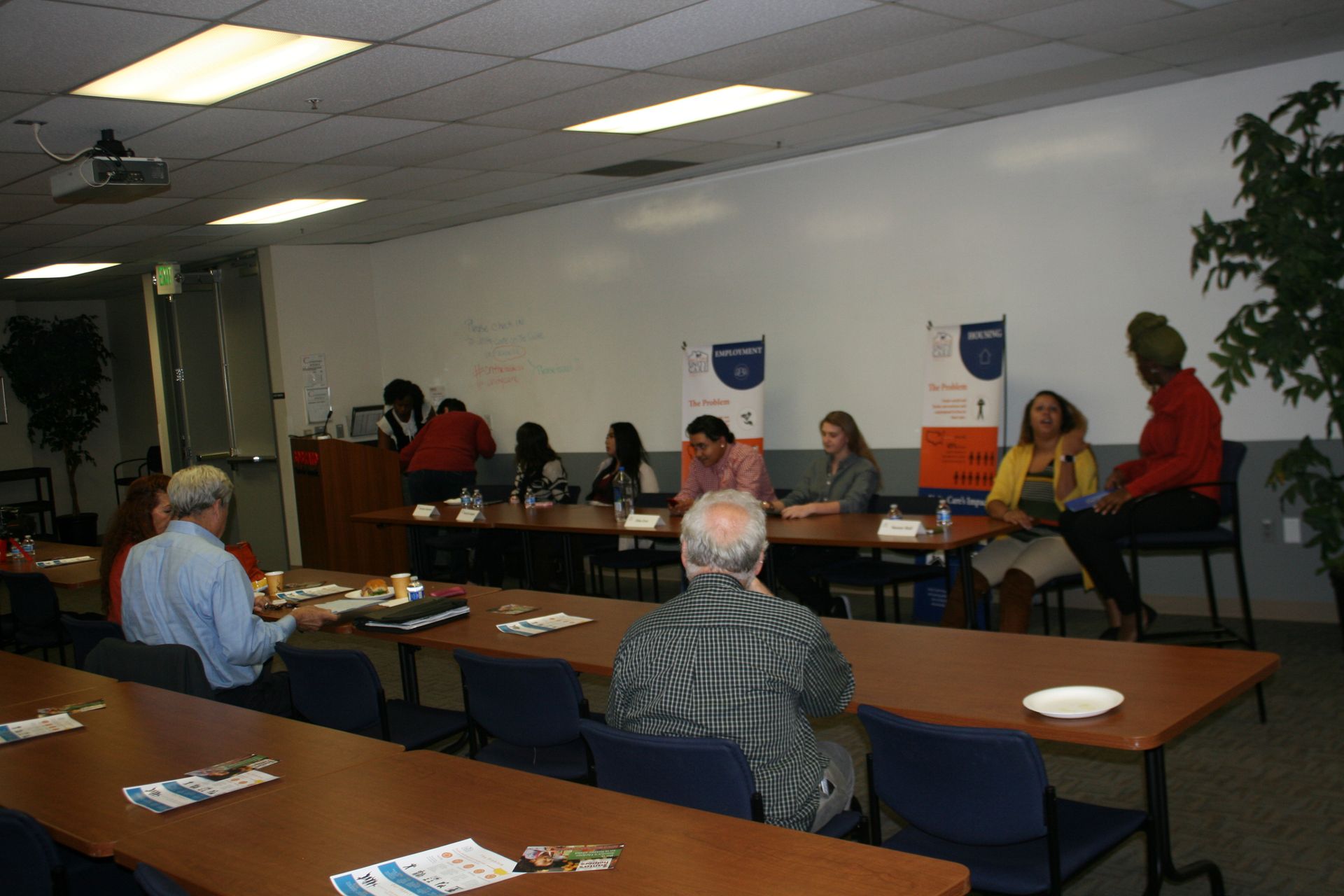 Meeting room with several people seated around tables, with a presenter at the front.