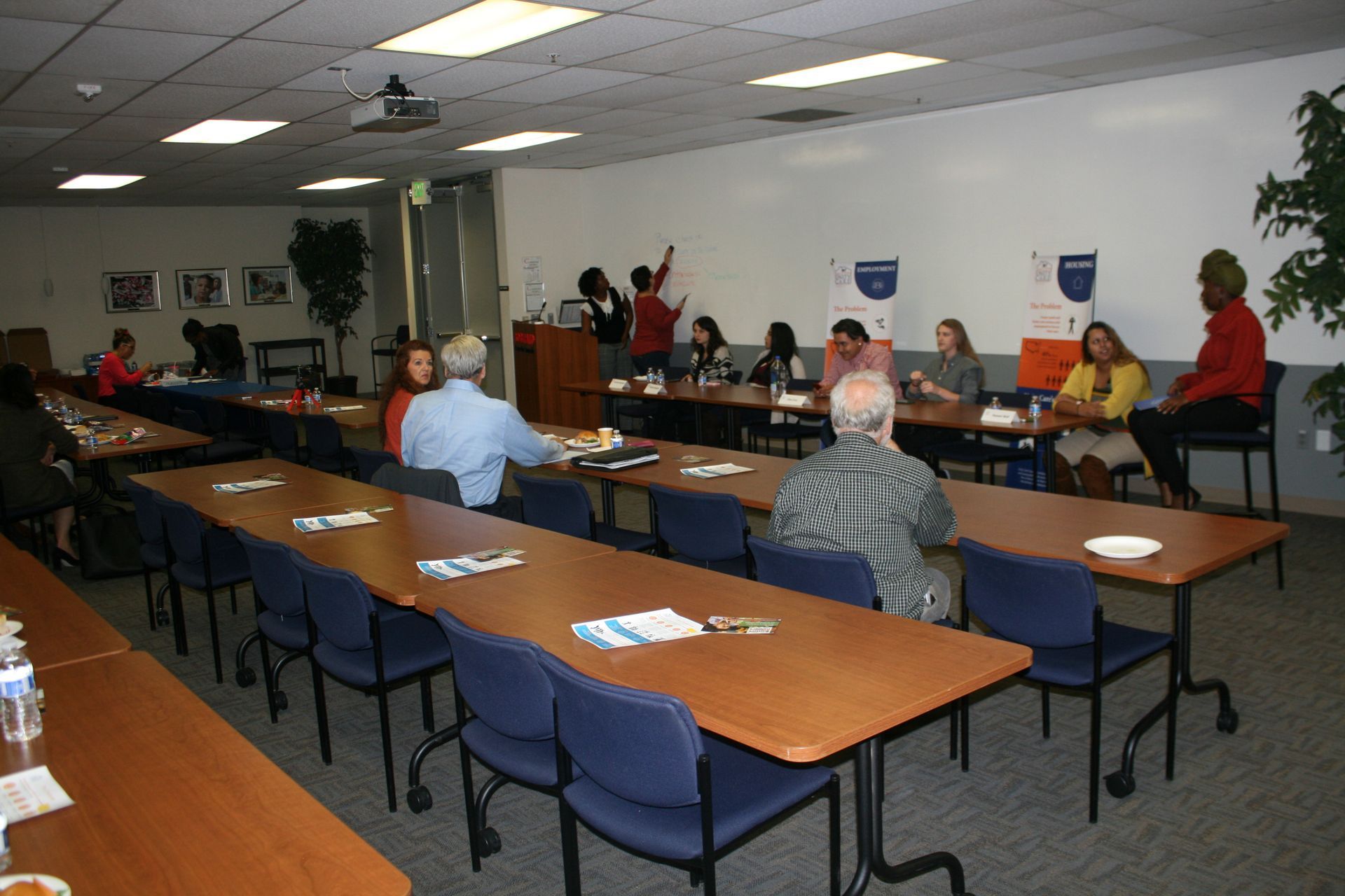 Meeting room with a few people seated around long tables, facing the front of the room