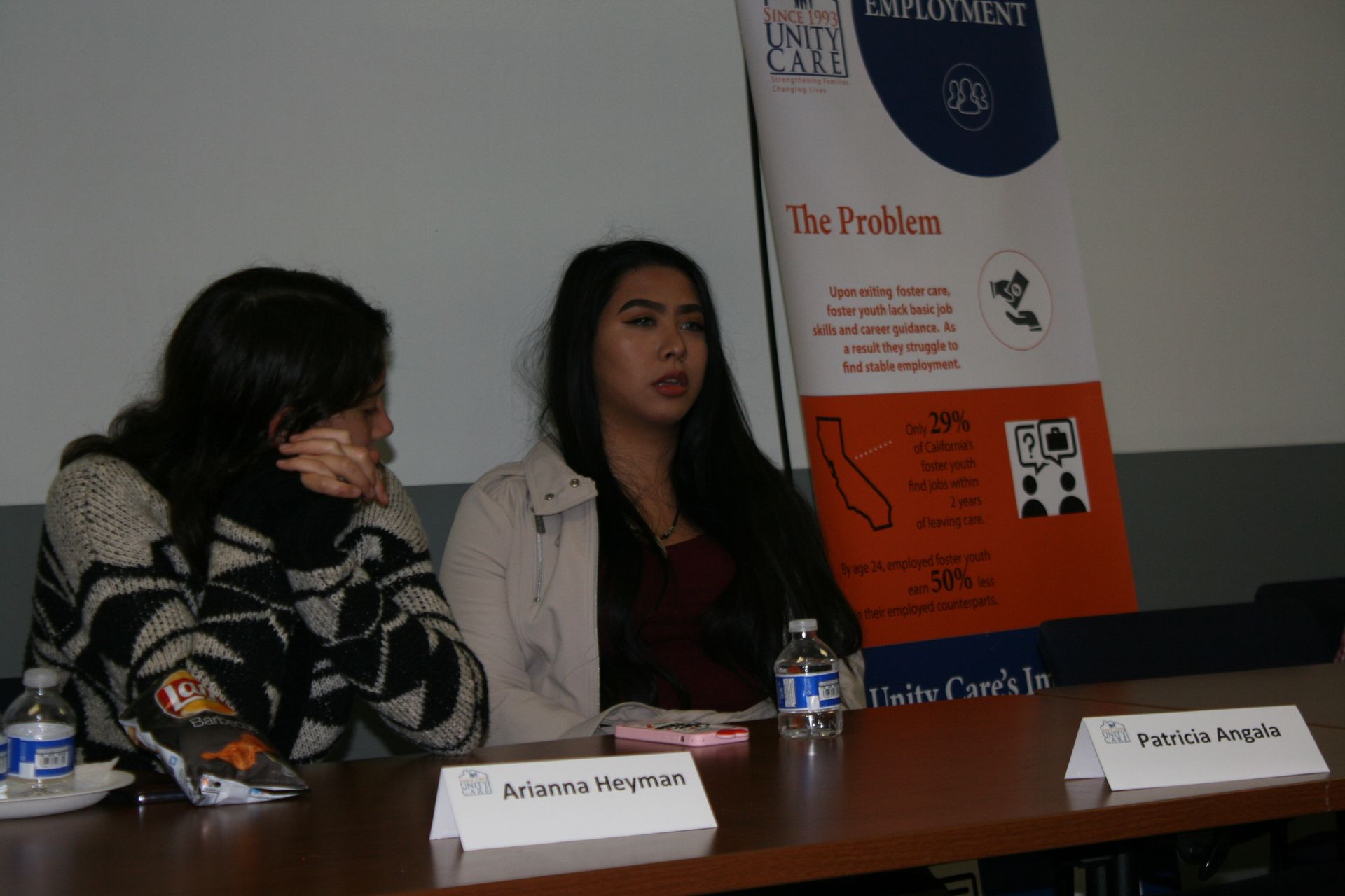 Two women seated at a conference table with name placards, speaking beside a poster presentation.