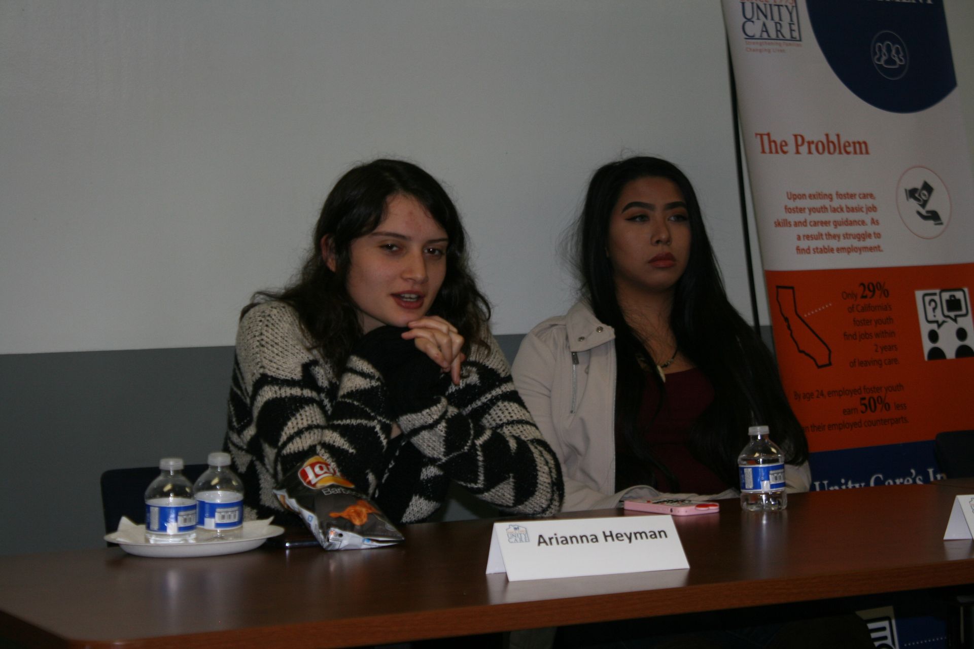 Two women seated at a panel table with microphones and water bottles, in front of a presentation poster.