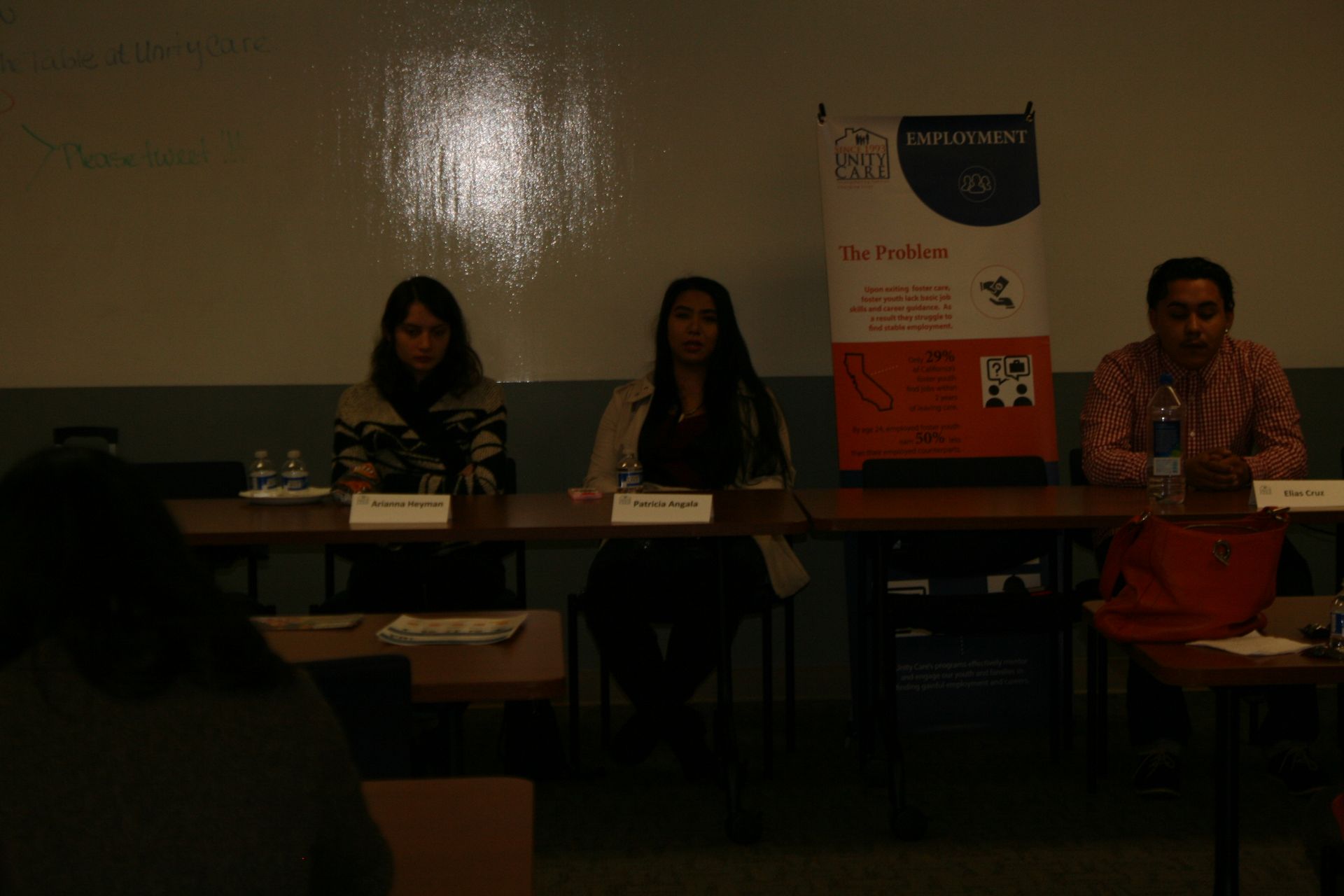Three people seated at a panel table in a dimly lit conference room, with a presentation poster behind them.