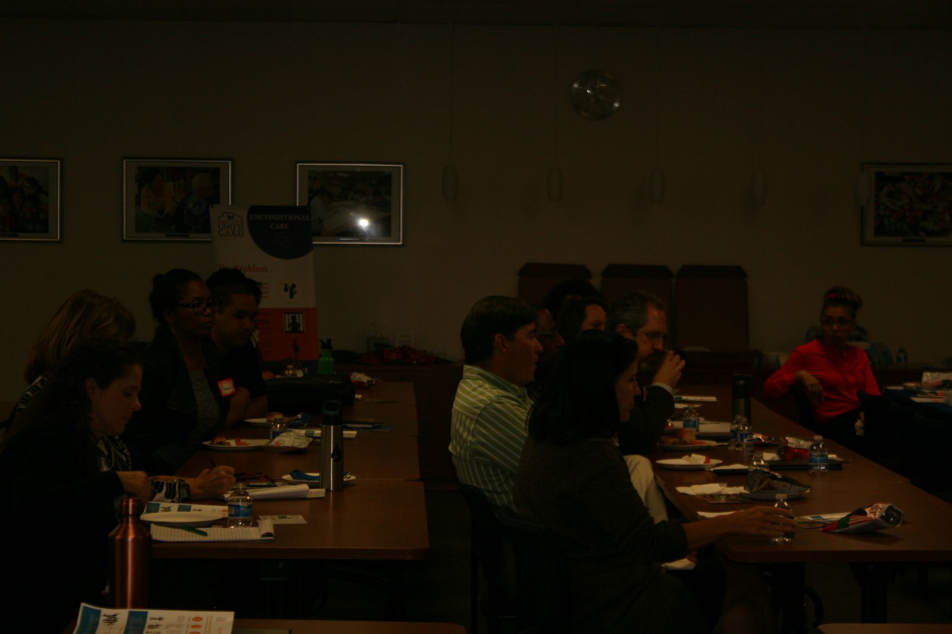 People seated at long tables in a dim meeting room, eating or talking during a gathering