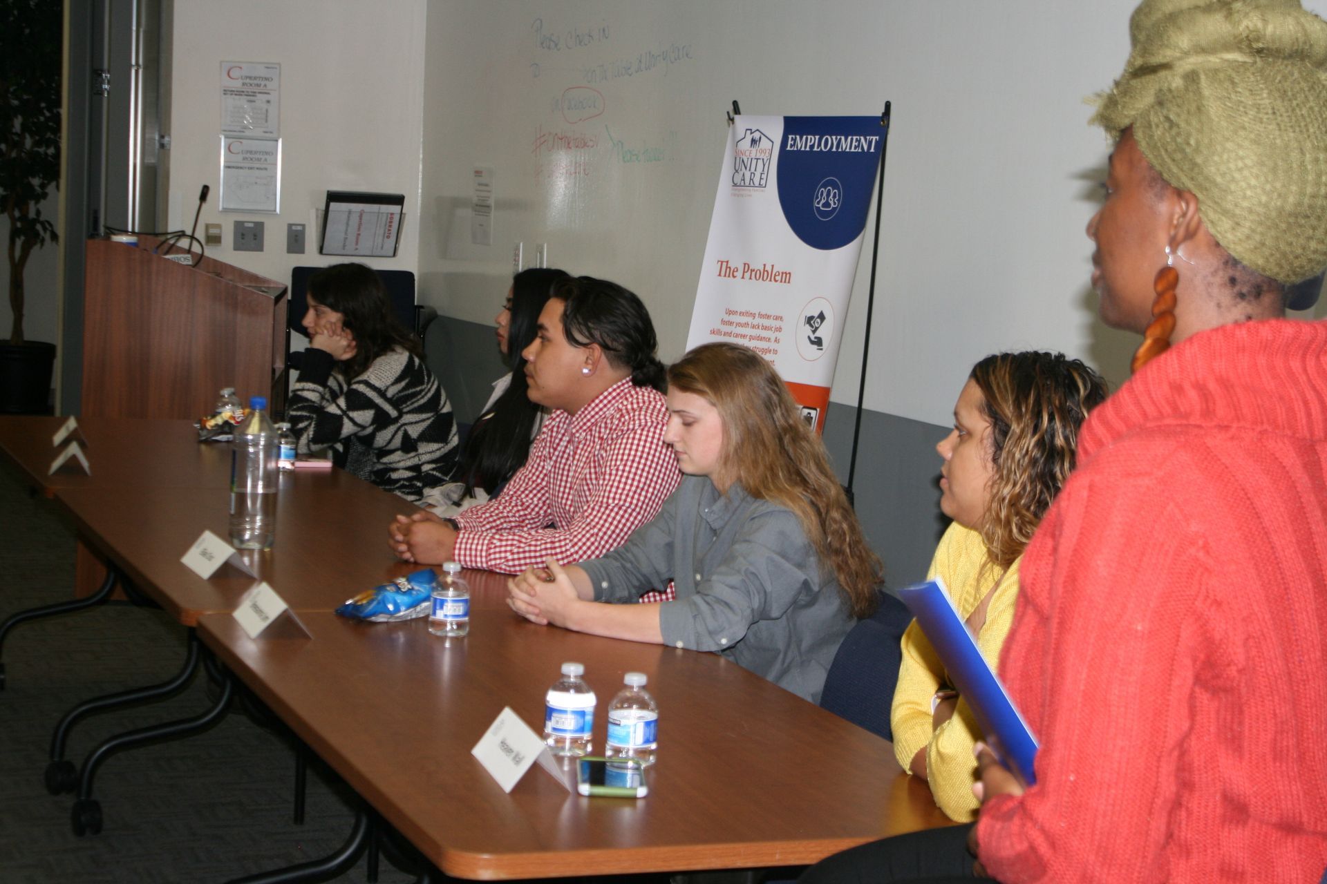 People seated at a conference table in a meeting room, listening to a speaker at right.