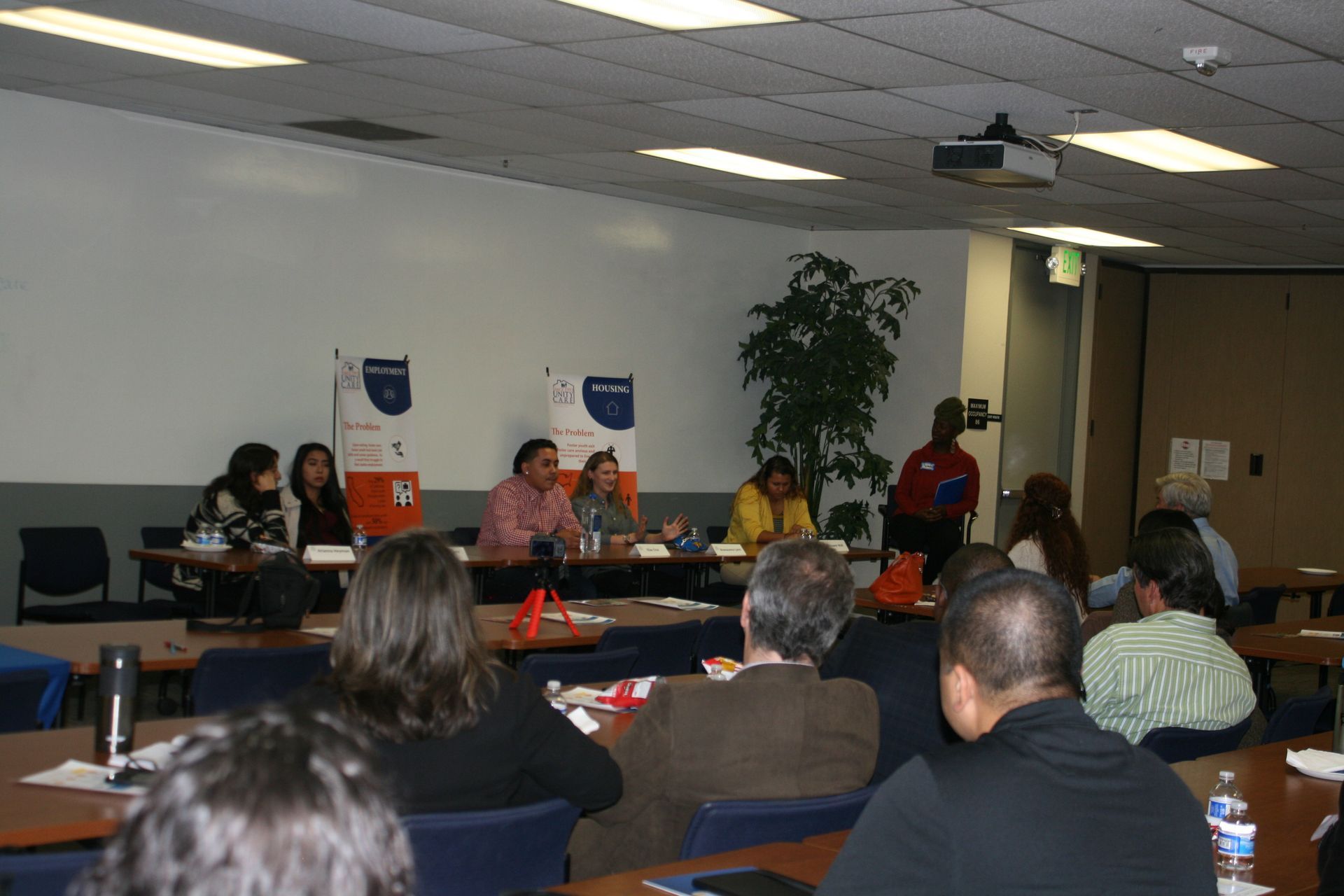 People seated in a classroom meeting, listening to speakers at the front.