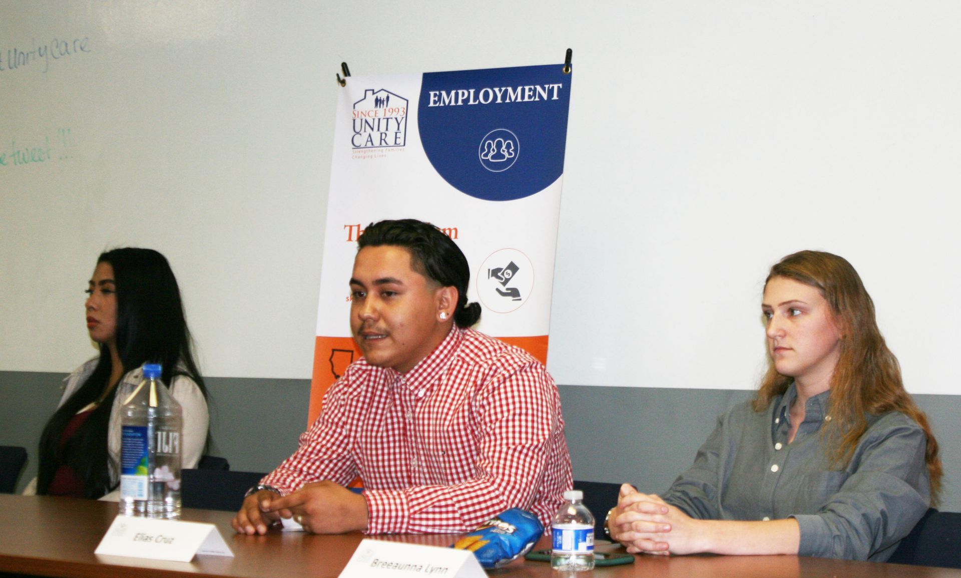 Panel discussion at a table, with a man in a red checkered shirt speaking between two women.