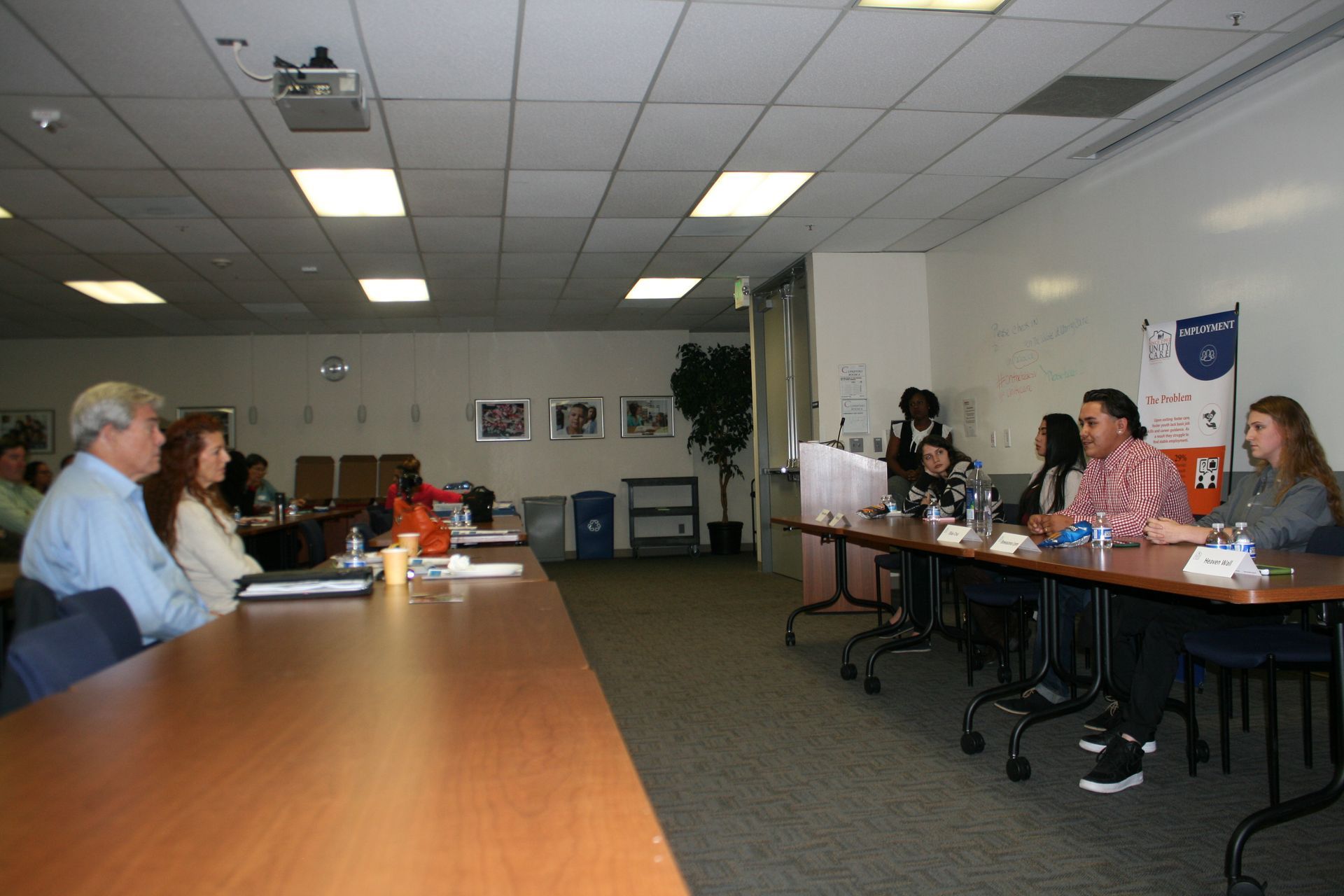 Conference room meeting with several people seated at tables, facing a presentation screen.