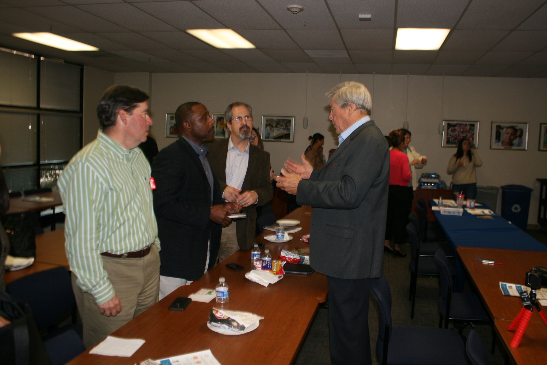 People chatting beside a conference table in a meeting room with papers, cups, and chairs.