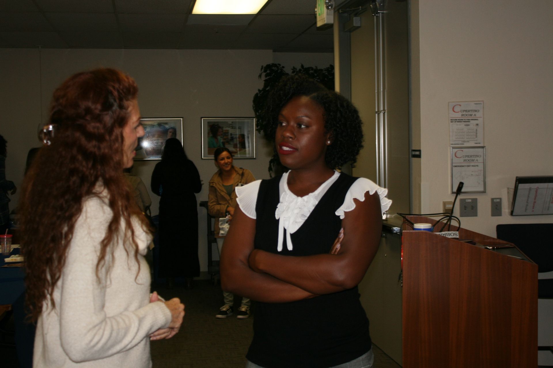 Two people talking in a hallway, one leaning against a podium with arms crossed.