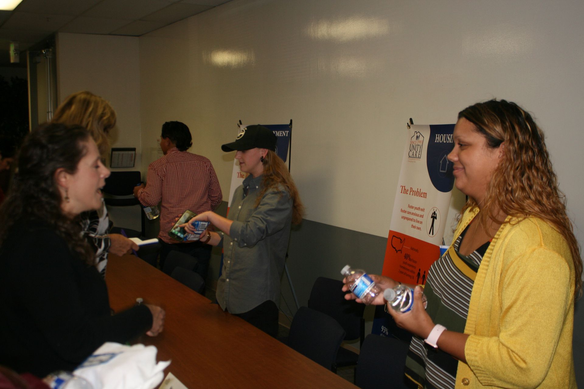 People exchanging items at a counter in a hallway, with a whiteboard and posters on the wall.