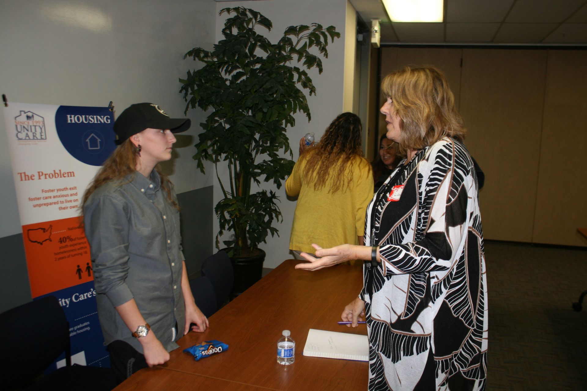 Two women talking at a table in a conference hallway, with a poster and plant in the background.