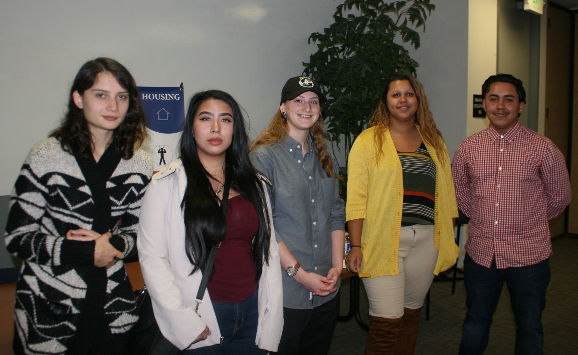 Five people standing indoors, posing for a group photo in front of a plain wall.