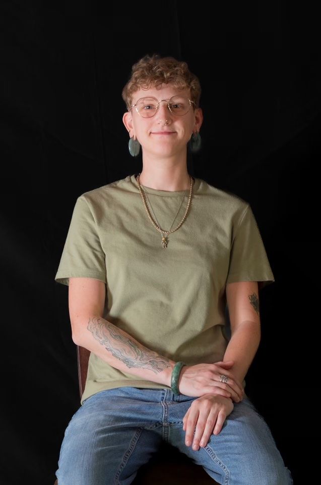 Person seated against a black background, wearing an olive T-shirt and jeans, with a calm expression.