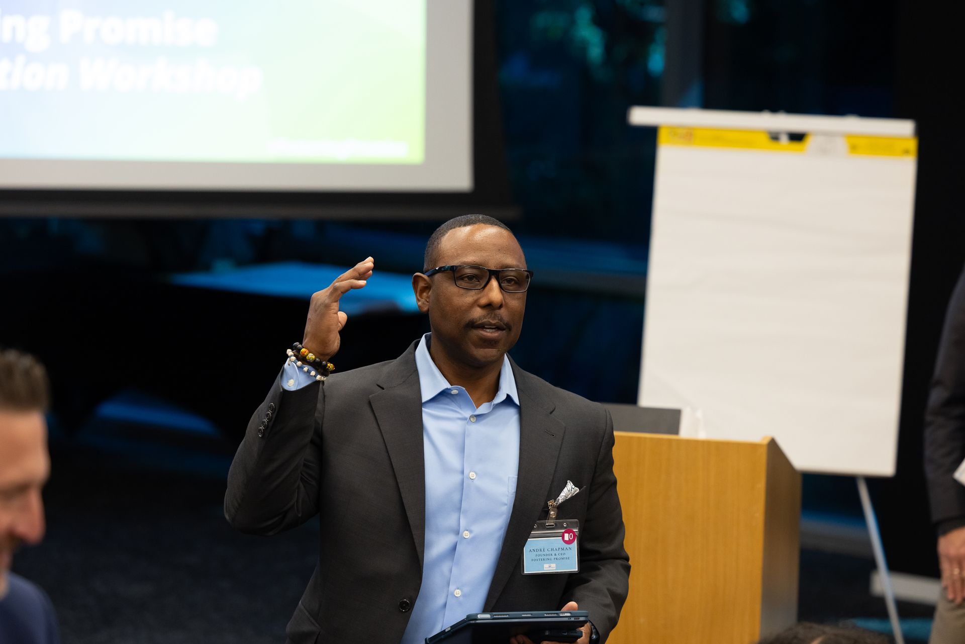 Speaker gesturing at a conference podium beside a flip chart and projected screen