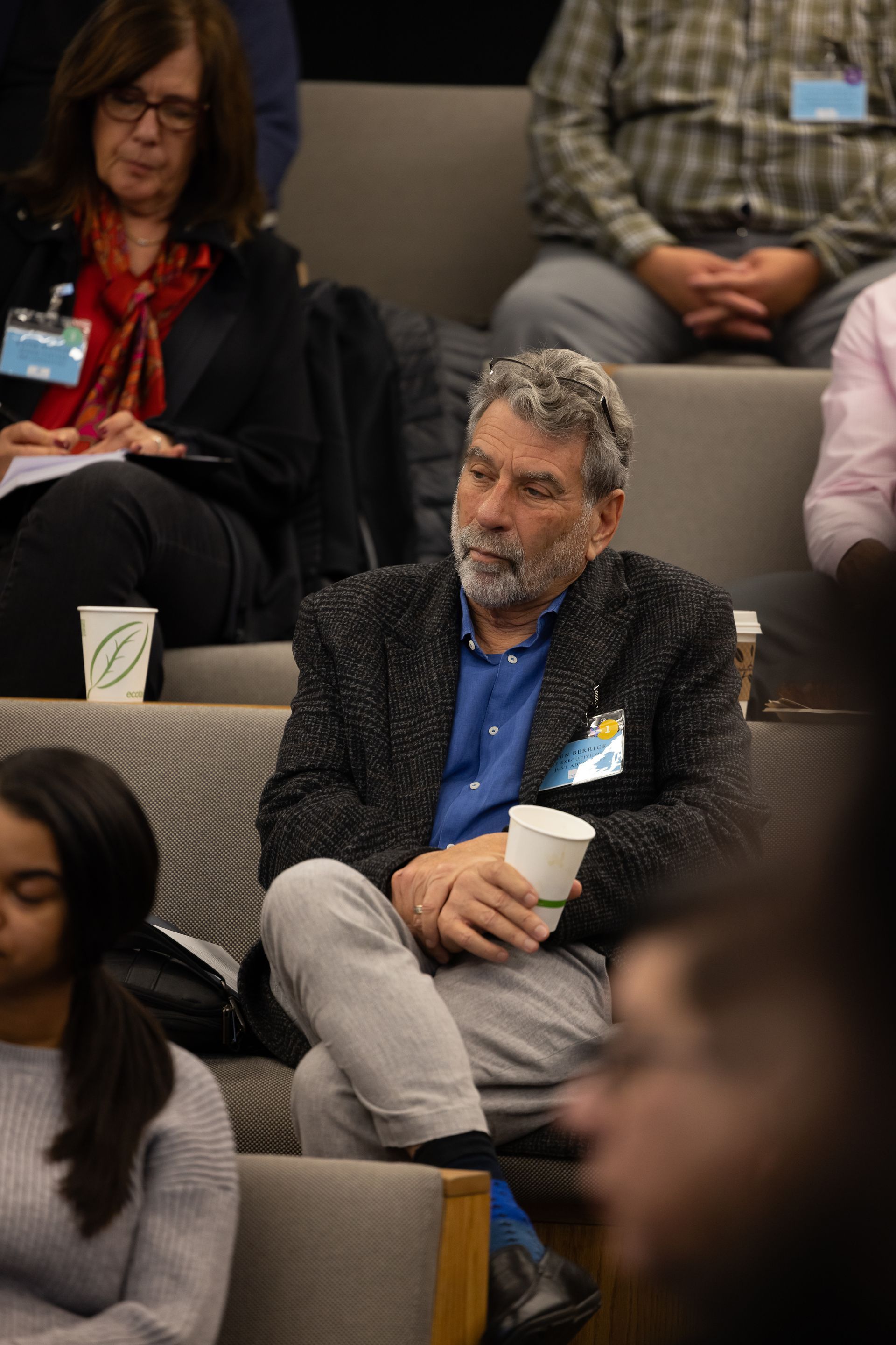 Audience member seated in a conference hall, wearing a blue shirt and dark jacket, looking thoughtfully ahead
