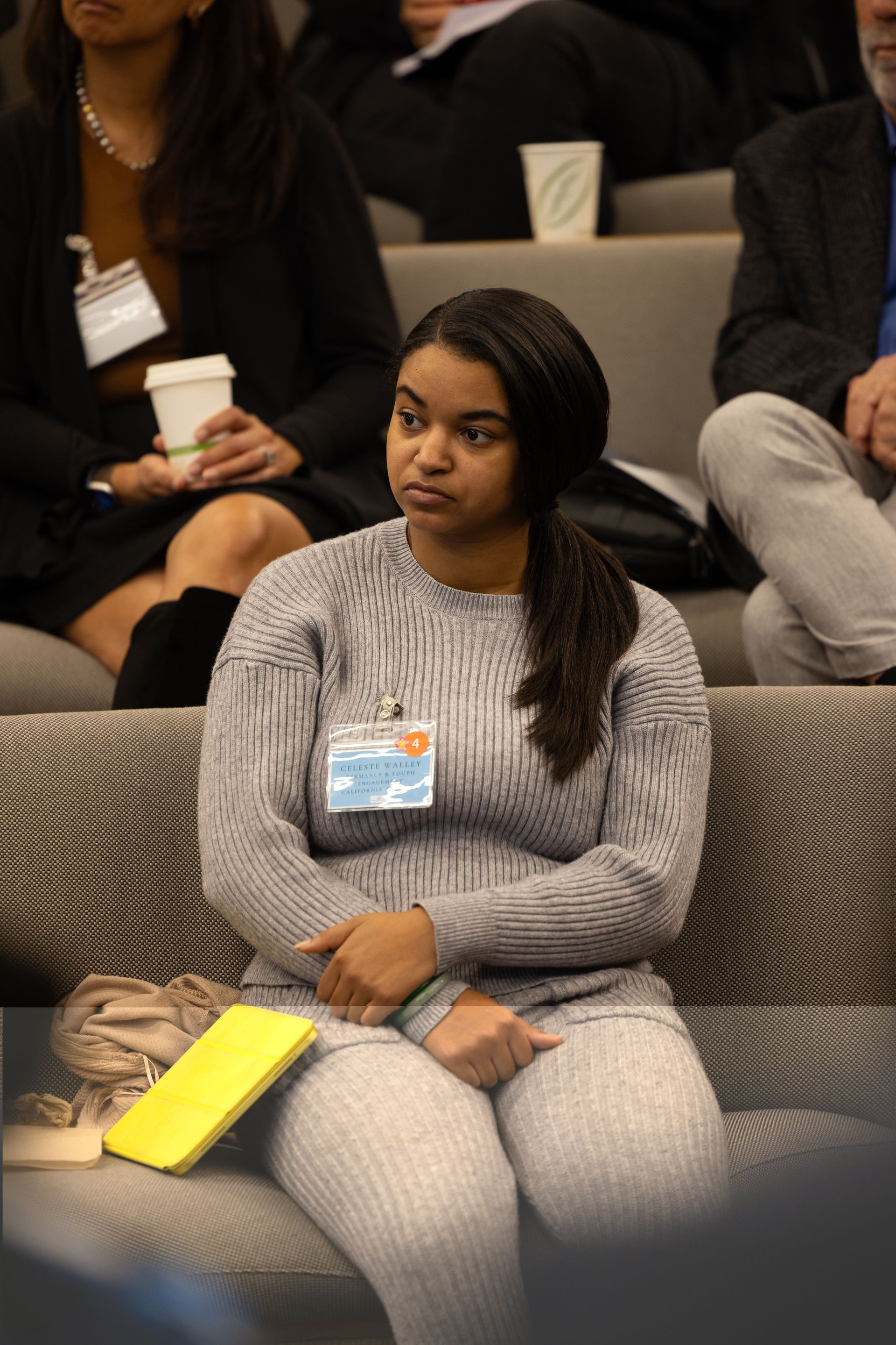Woman in a gray sweater sitting on a couch at a conference, wearing a name badge and looking to the side.