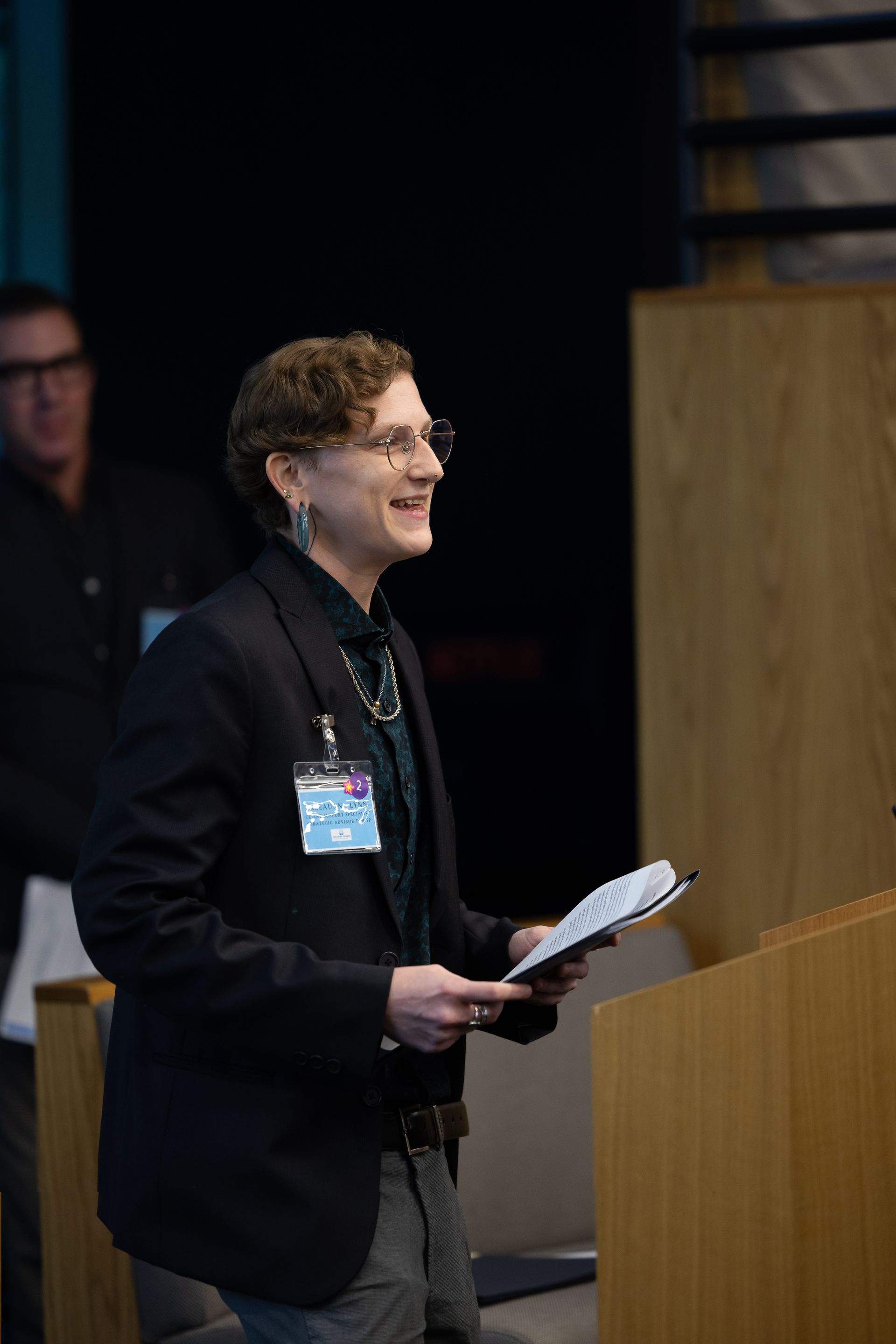 Person speaking at a podium, holding papers and smiling in a dimly lit conference room.