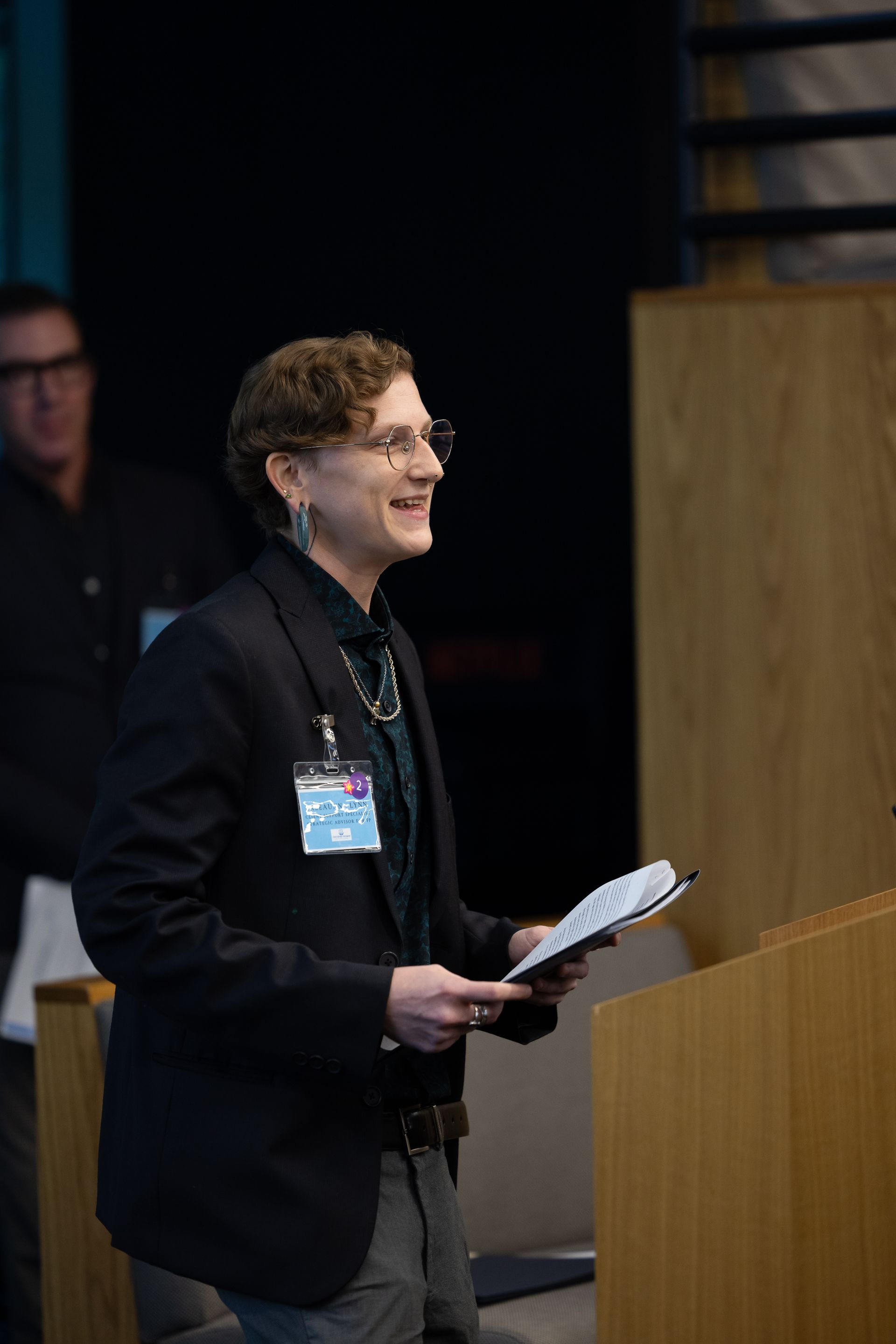 Person speaking at a podium, holding notes and smiling in a dim conference room.