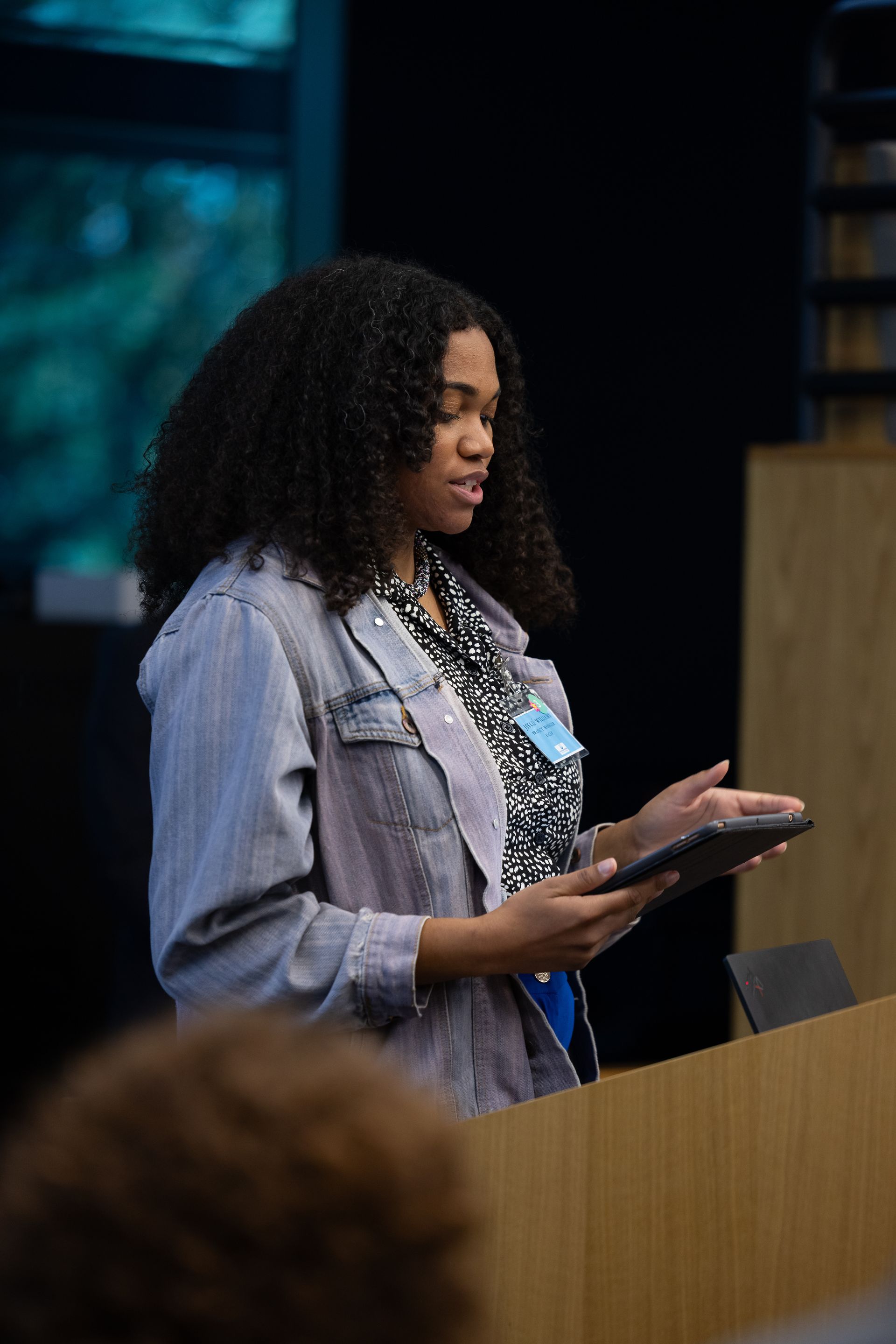 Woman speaking at a podium, holding a tablet, in a conference setting