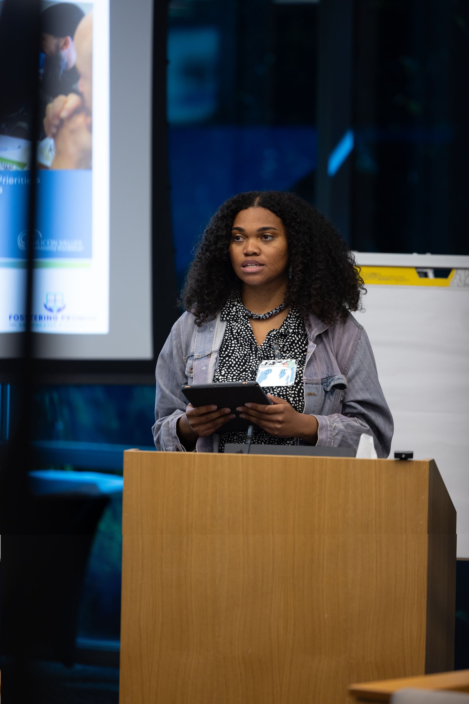 Woman speaking at a podium indoors, holding a tablet, with a presentation screen behind her.