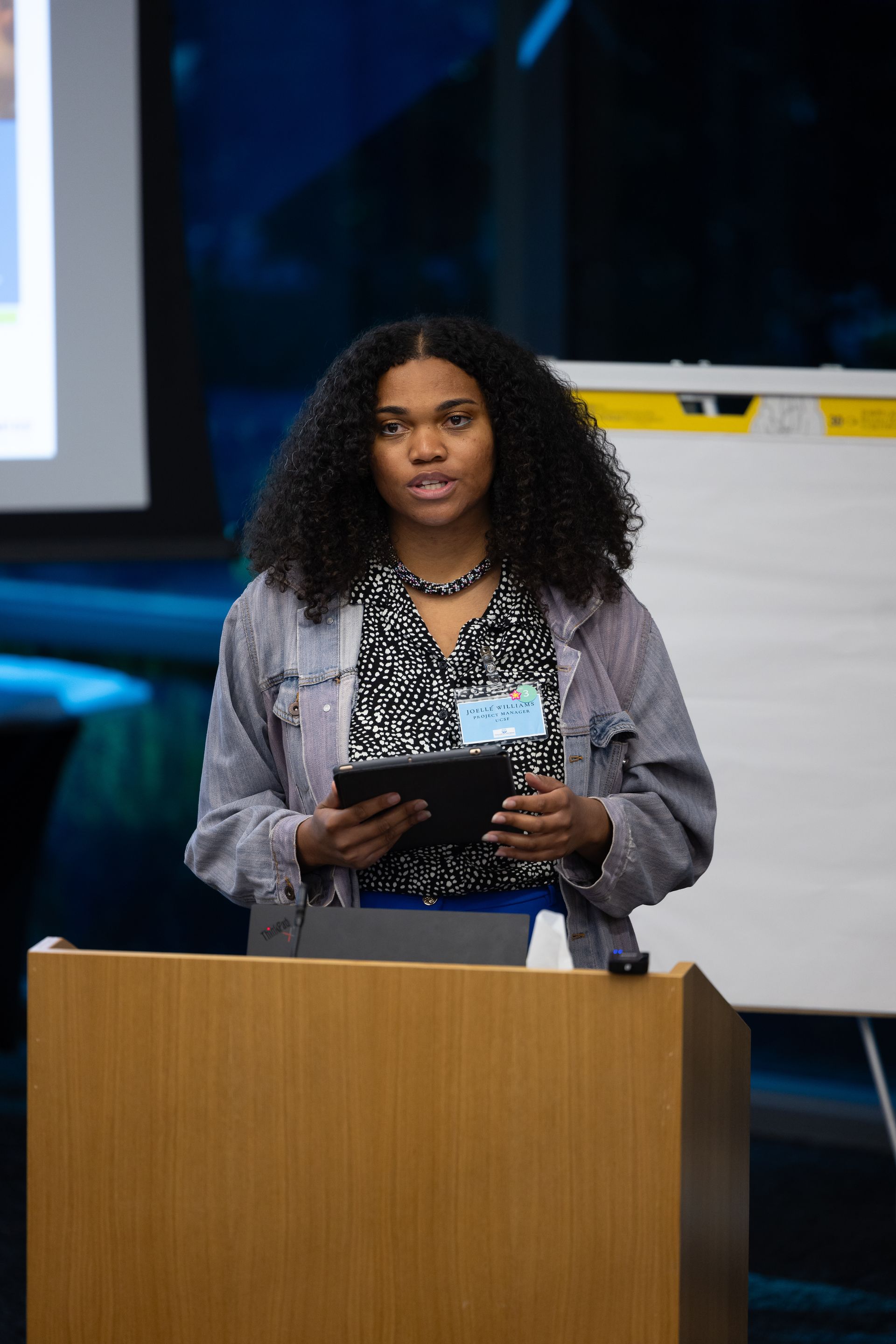 Woman speaking at a podium, holding notes in a conference setting.