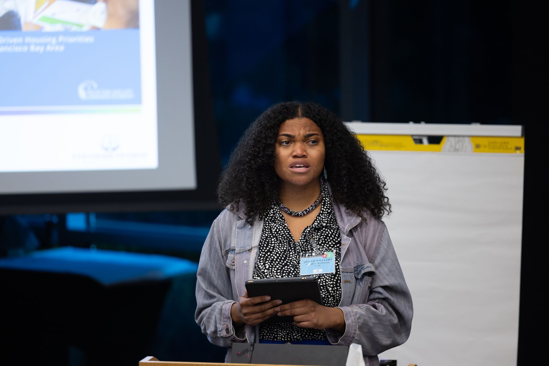 Woman speaking at a podium during a presentation, holding notes in front of a projected screen.