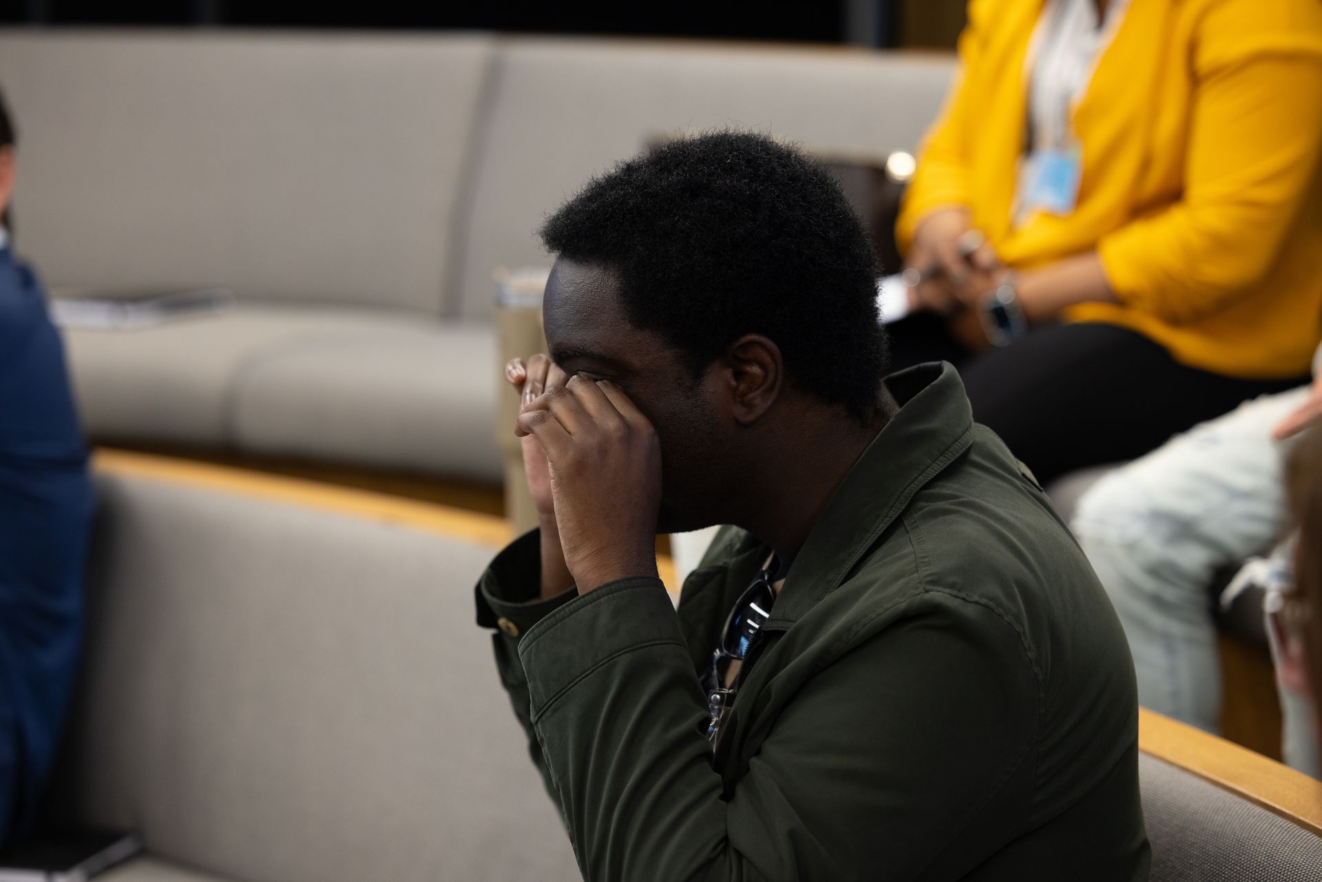 Person seated indoors, hands covering face, with others blurred in the background.