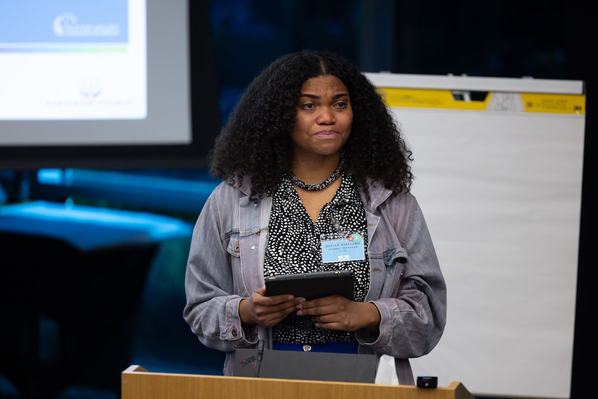 Speaker at podium holding notes during a presentation in a conference room