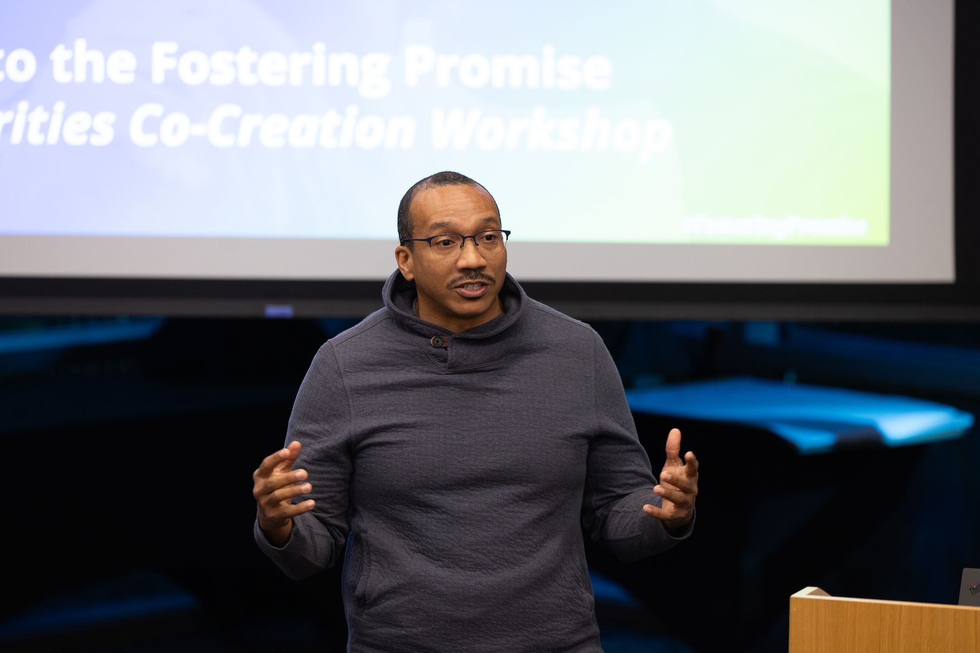 Speaker presenting on stage in a dark sweater, gesturing in front of a projected slide.