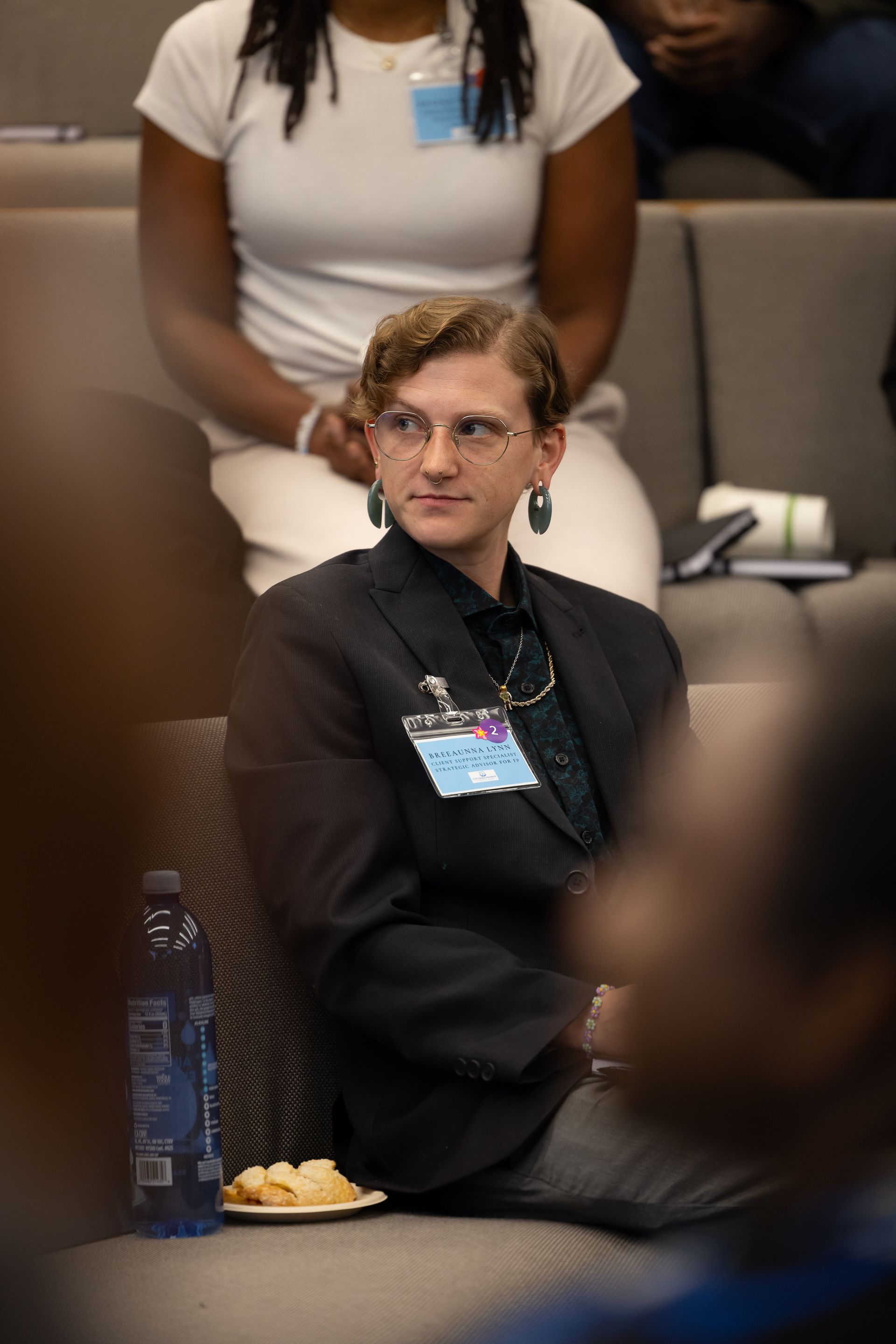 Audience member seated indoors, wearing a black blazer and glasses, looking to the side with a snack and water bottle nearby