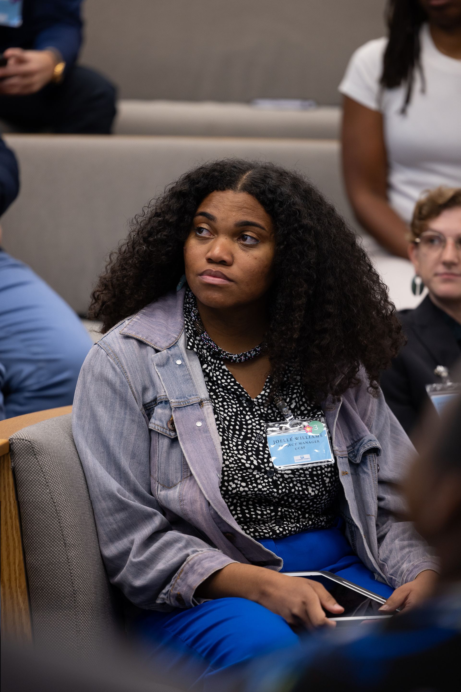 Audience member seated in a lecture hall, wearing a gray blazer and blue pants, looking to the side.