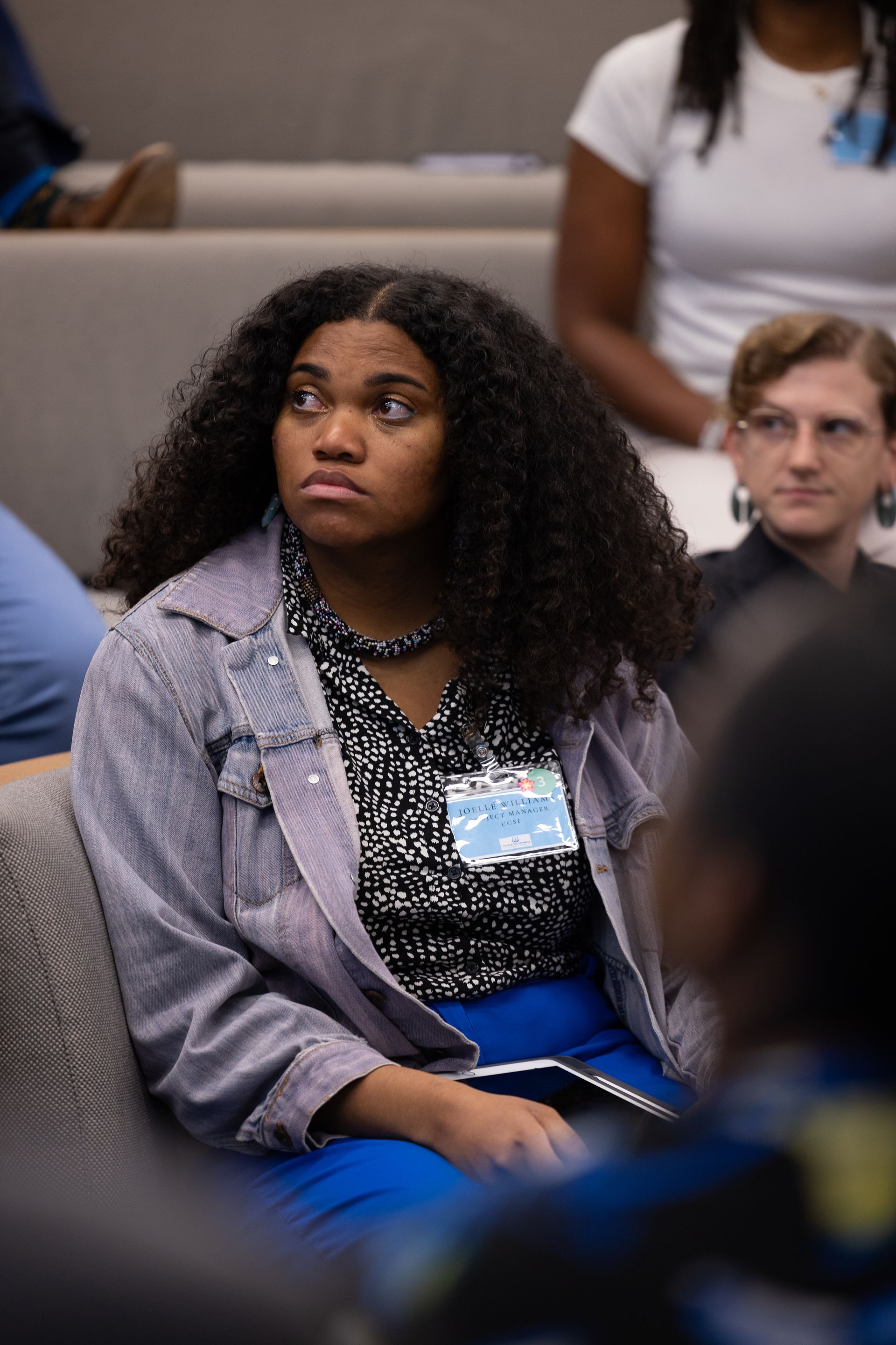 Audience member seated in a lecture hall, wearing a gray jacket and blue skirt, looking attentively to the side