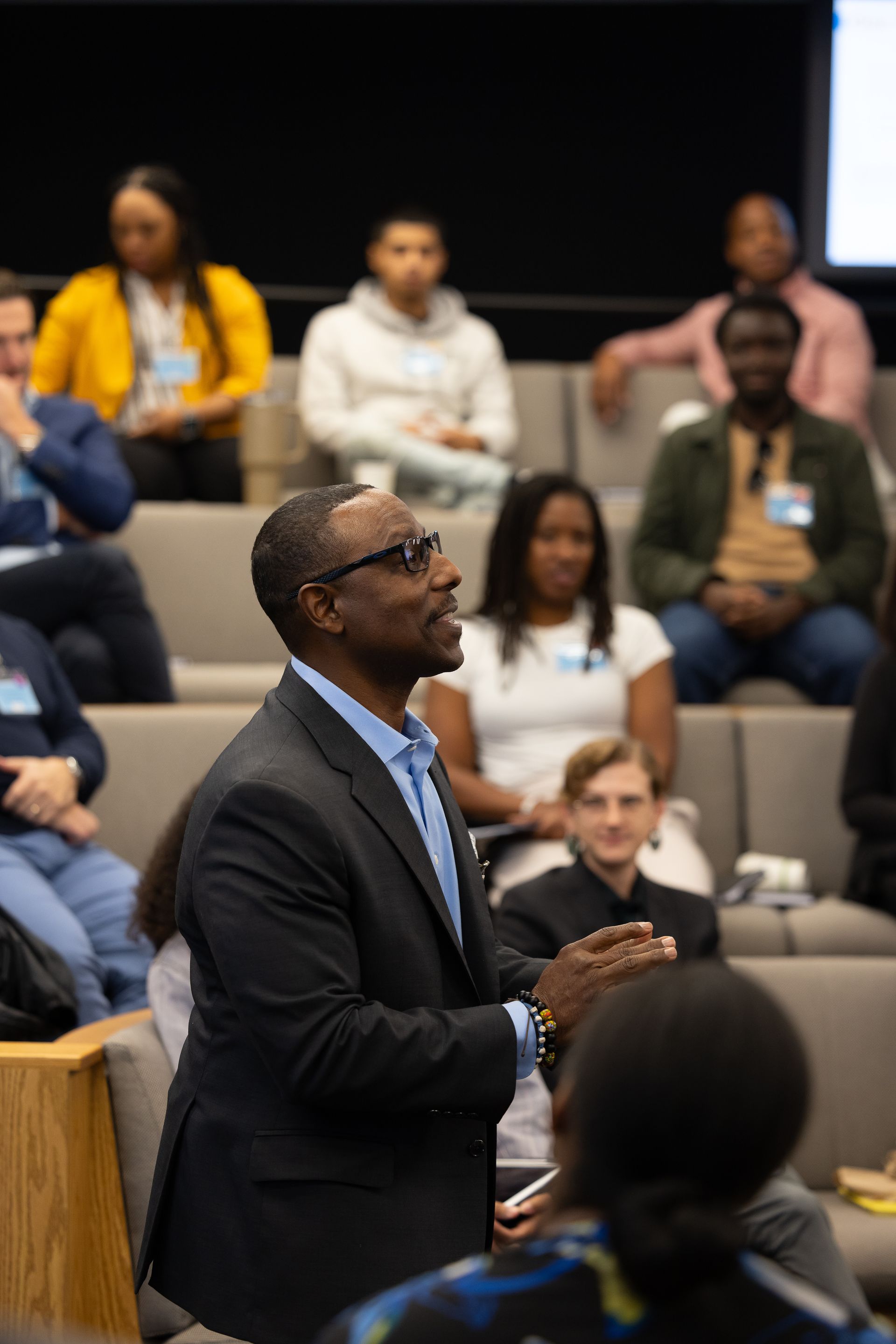 Speaker addressing an audience in a lecture hall, wearing a dark blazer and glasses, with seated attendees behind him