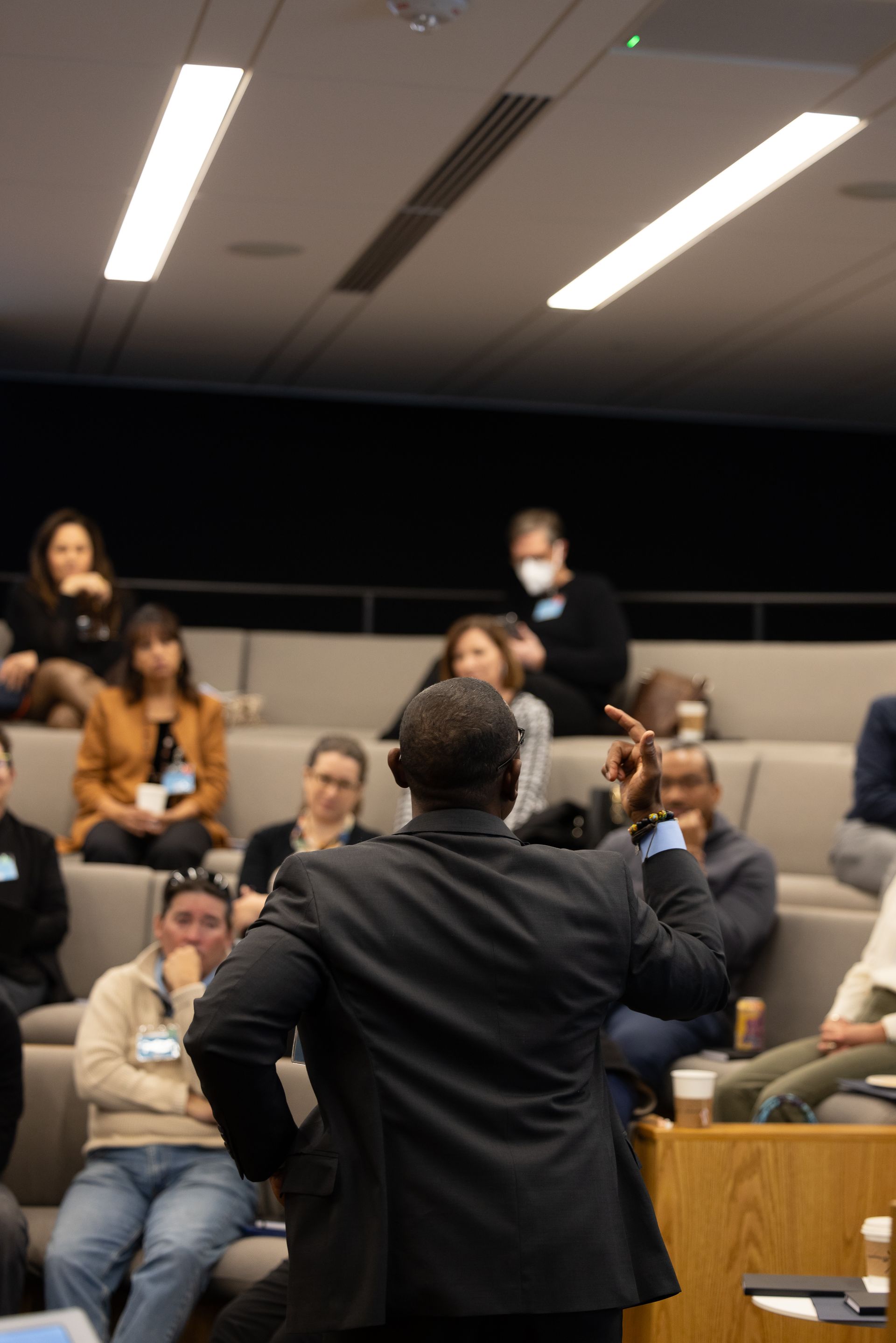 Speaker addressing seated audience in a classroom or lecture hall, with overhead lights above.