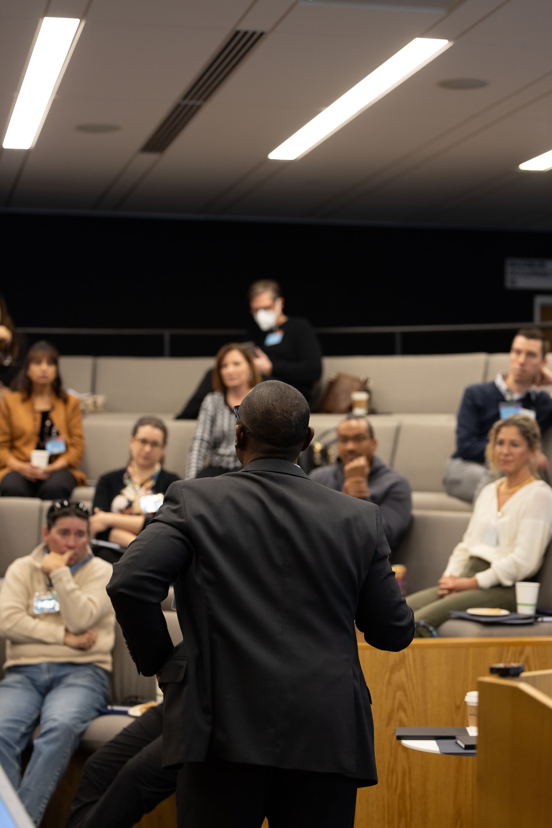Speaker addressing seated audience in a lecture hall, viewed from behind.
