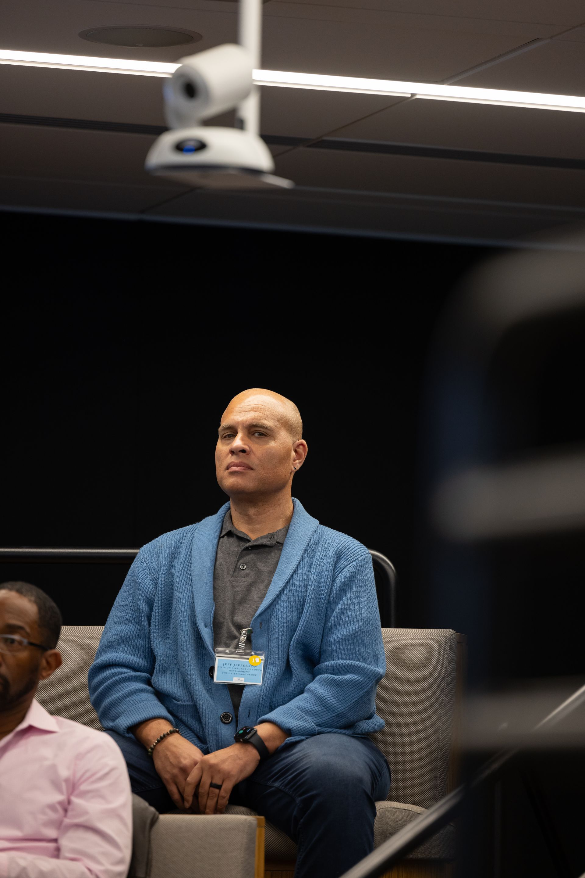 Seated man in a blue jacket at a conference, with a ceiling camera overhead