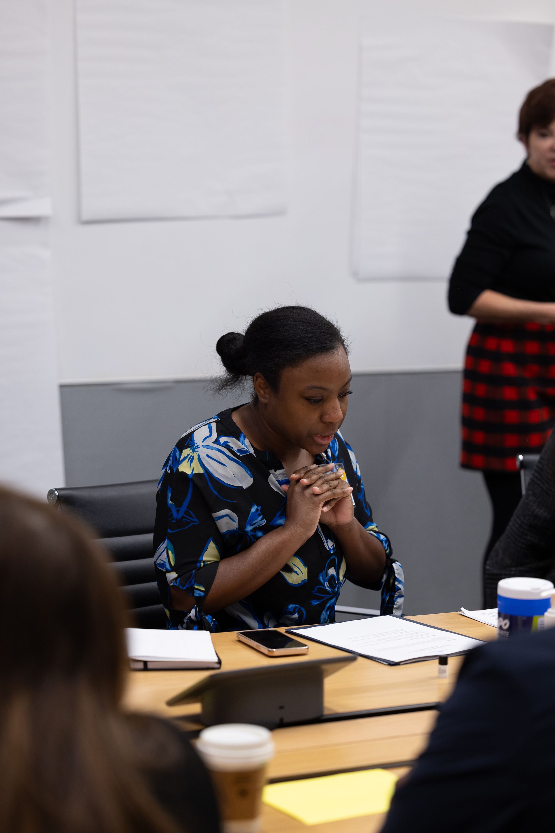 Woman speaking at a desk in a meeting room, with papers and a coffee cup nearby.