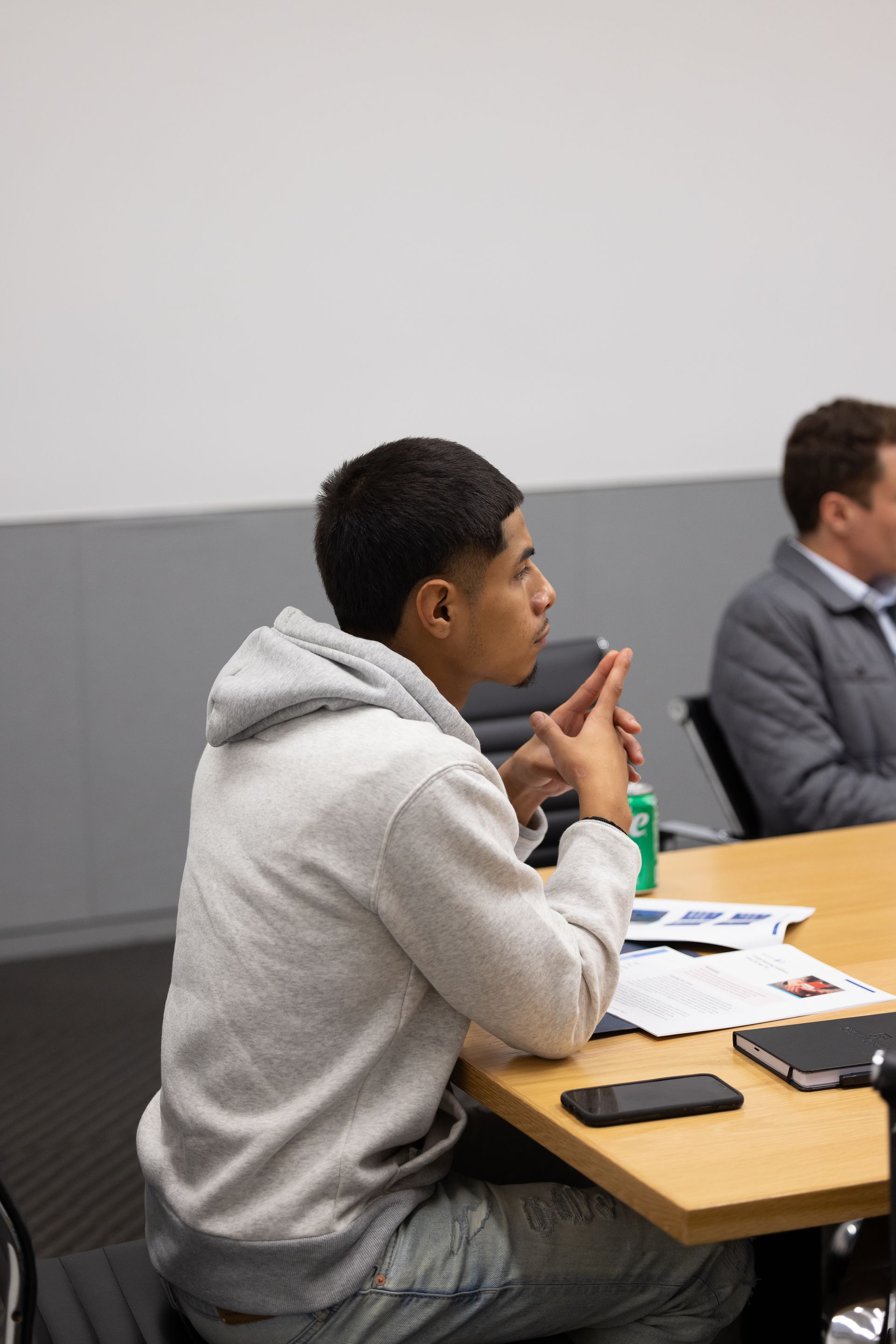 Person in a gray hoodie sits at a table in a meeting room, listening attentively with papers and a phone nearby.