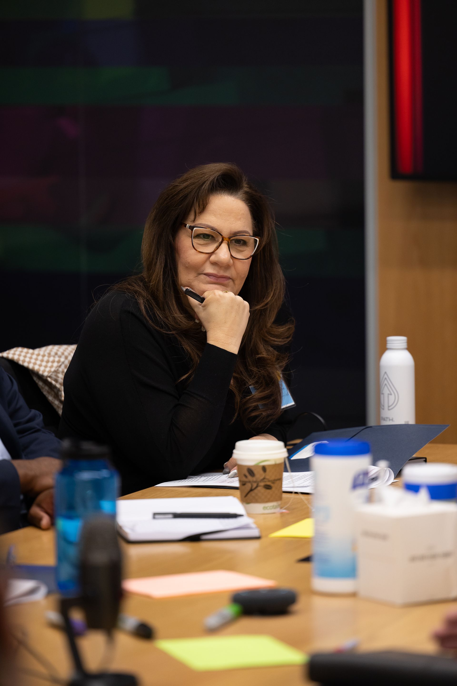 Woman seated at a conference table, resting her chin on her hand, with coffee cups and papers nearby.