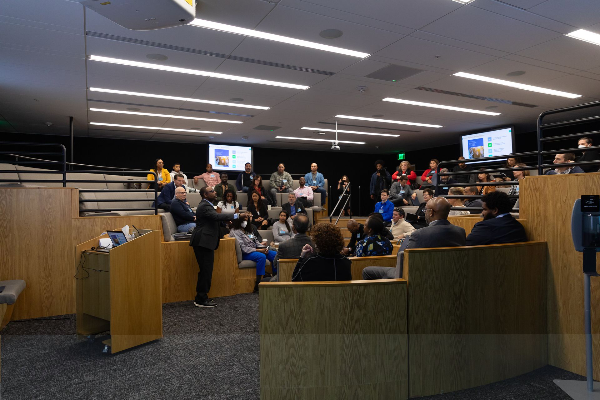 Auditorium meeting with seated audience and presenters at the front under bright ceiling lights.