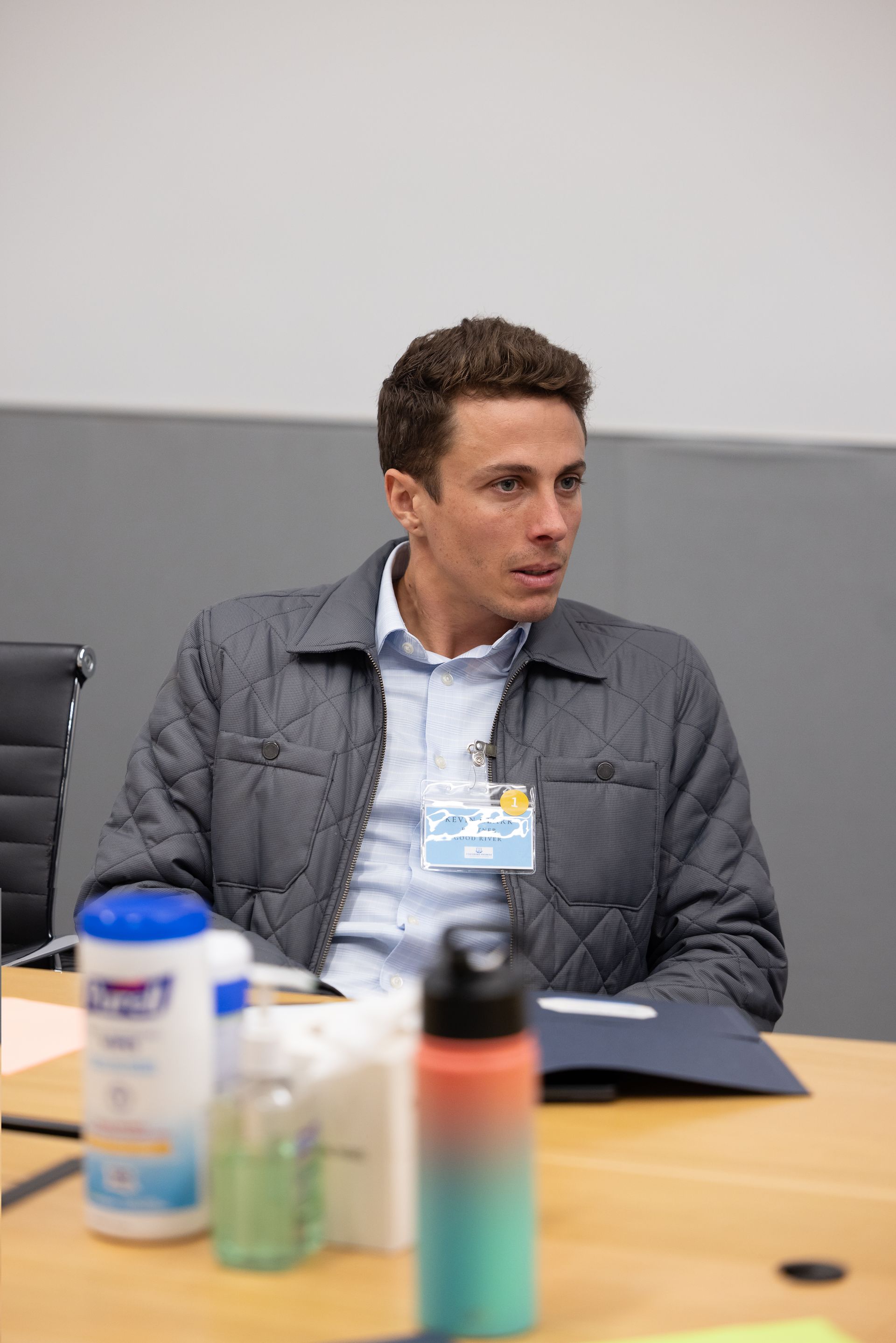 Man in a gray quilted jacket seated at a conference table with drinks in the foreground, looking to the side