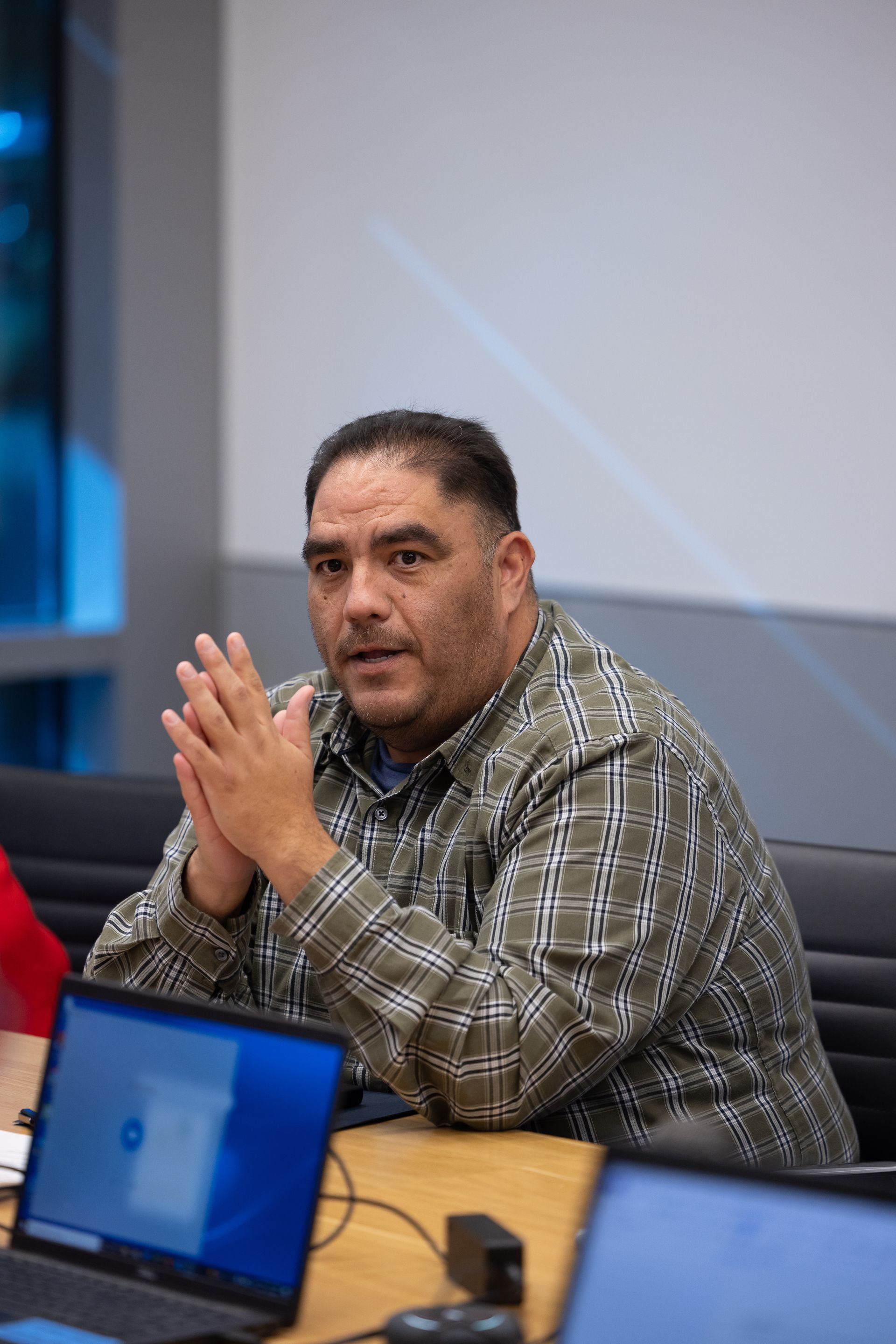 Man sitting at a conference table, speaking with hands clasped, in a meeting room with laptops nearby.